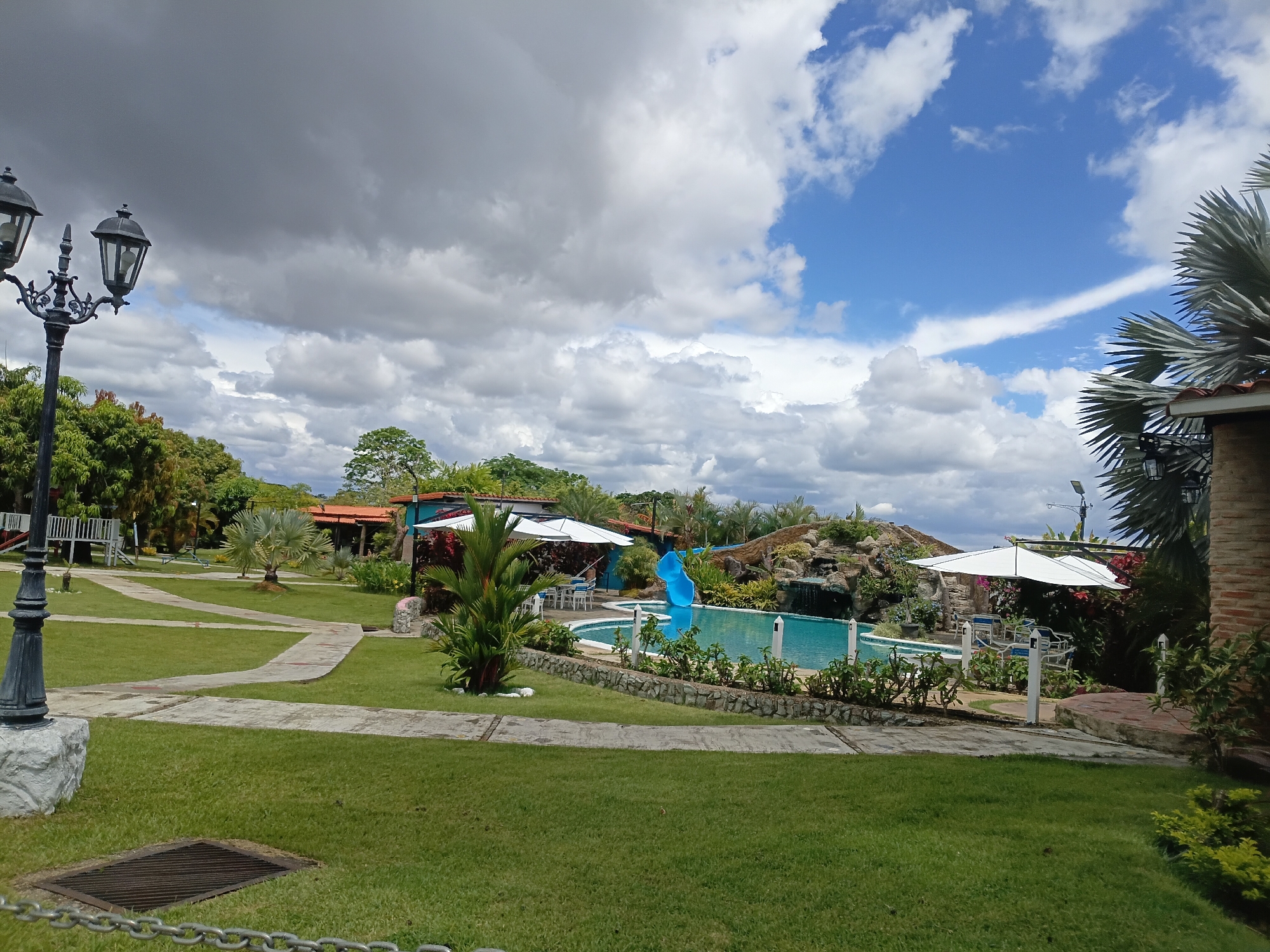 This image presents a serene outdoor recreational area, likely a resort or large private estate, under a strikingly dynamic sky. The scene is characterized by lush green lawns and abundant tropical vegetation, indicative of a warm climate.
In the foreground, a decorative black lamppost with two lanterns stands on a white stone base to the left. A metal grate is visible on the grass, along with a metal chain. Concrete paths, winding and irregularly shaped, traverse the manicured lawn.
The midground features a sparkling blue, kidney-shaped swimming pool. A bright blue water slide empties into the pool from a rocky, landscaped area that includes a small waterfall. White outdoor umbrellas provide shade over seating arrangements around the pool, which is bordered by low green bushes and white decorative posts. Various palm trees and vibrant tropical plants are scattered throughout the landscape. Buildings with reddish roofs are partially visible among the trees in the background, along with what appears to be a wooden structure or play area to the far left.
The sky is a notable element, displaying a dramatic contrast. The left side is dominated by heavy, dark grey storm clouds, suggesting imminent rain or recent stormy weather. Conversely, the right side transitions to a clear, brilliant blue sky adorned with fluffy white cumulus clouds, indicating sunny conditions. This creates a visually arresting split across the heavens.
The time of day appears to be daytime, possibly late morning or early afternoon, with natural lighting illuminating the scene. No people are visible, and there is no discernible text in the image. The overall impression is one of a tranquil and well-maintained leisure space.