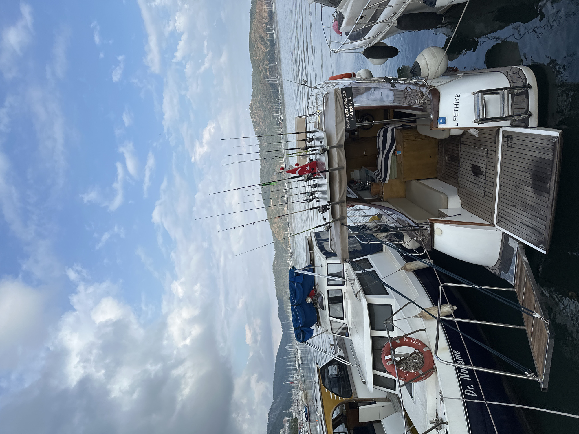 A motor yacht is docked in Fethiye, Turkey. The yacht is white and navy blue, with fishing rods along the top. A Turkish flag is visible. The text on the yacht reads "Dr. Nolame". A life preserver is also visible. In the background, there is a hilly coastline, and other boats are docked nearby. The sky is partly cloudy.