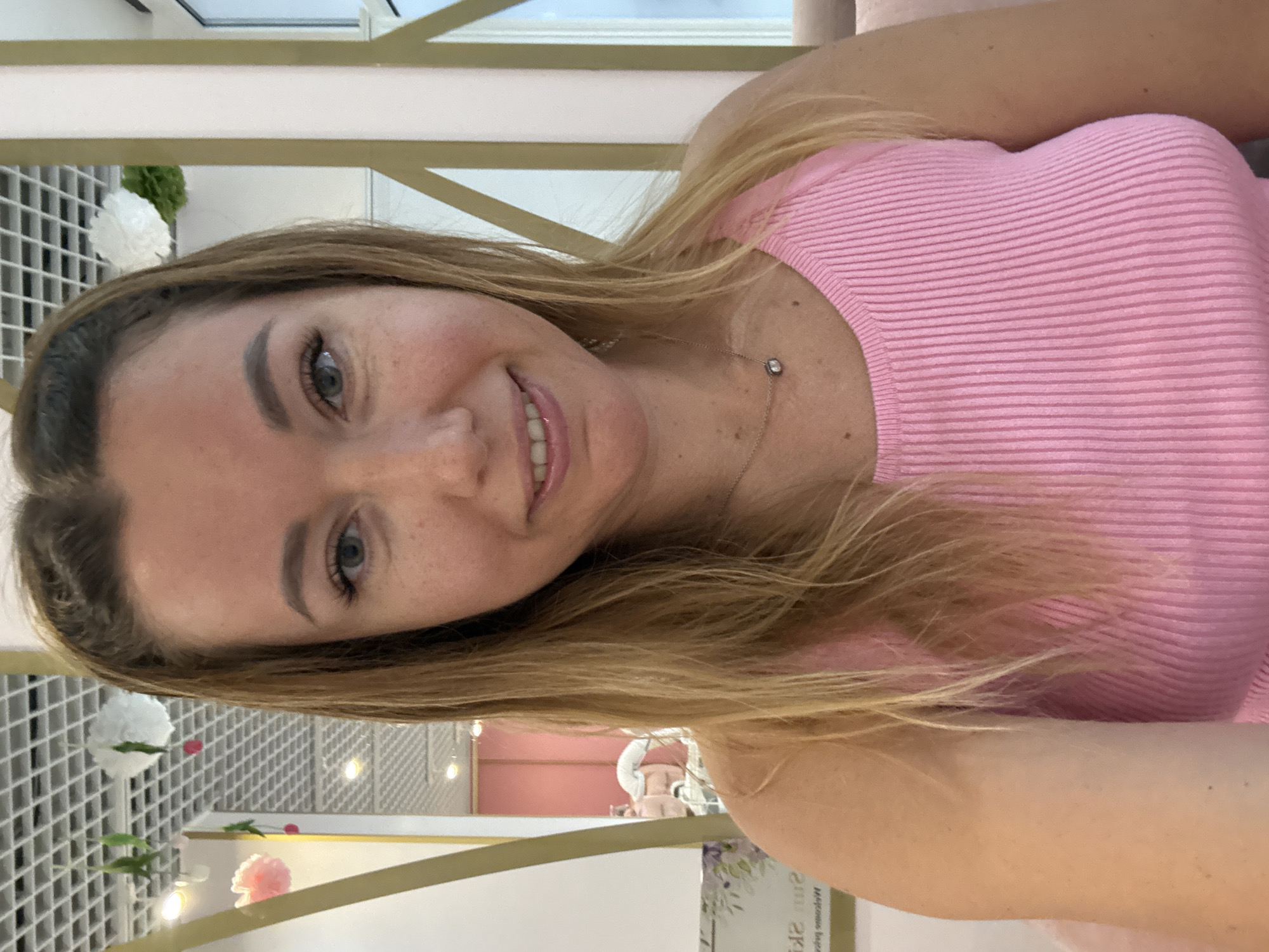 An indoor, medium shot shows a Caucasian woman with blonde hair and light blue eyes, looking directly at the camera. She is wearing a pink ribbed tank top and a silver necklace with a small circular pendant. Her hair is pulled back from her face, with some strands framing her face. She has freckles across her nose and cheeks. The background appears to be a salon or spa, with a white drop ceiling with a grid pattern, some decorative pink and white pom-poms hanging from the ceiling, and gold and pink accents on the walls. A mirror in the background reflects a pink wall and a person's head and shoulders. The lighting is bright and even.