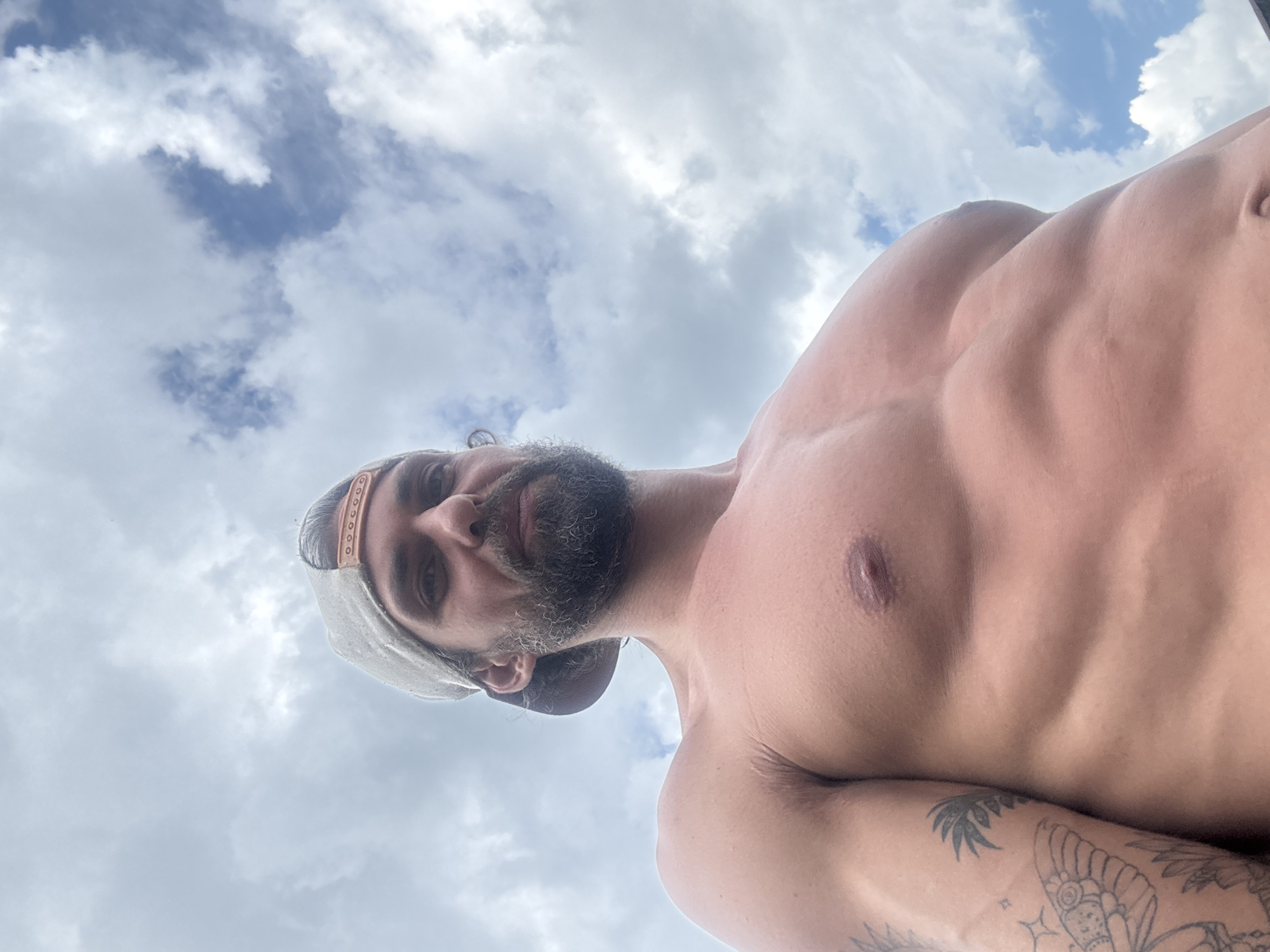This media file shows a close-up, low-angle, upside-down shot of a man's torso and face against a cloudy sky. The man is shirtless and has well-defined abdominal muscles and some tattoos on his arm, including what appears to be a butterfly design. He is wearing a baseball cap. His face is partially visible, with a beard and intense eyes looking directly at the camera. The lighting suggests it is daytime, and the sky is filled with both white, fluffy clouds and patches of blue, indicating variable weather. There are no visible text elements in the image. The context provided indicates the location is Kuti, Russia.