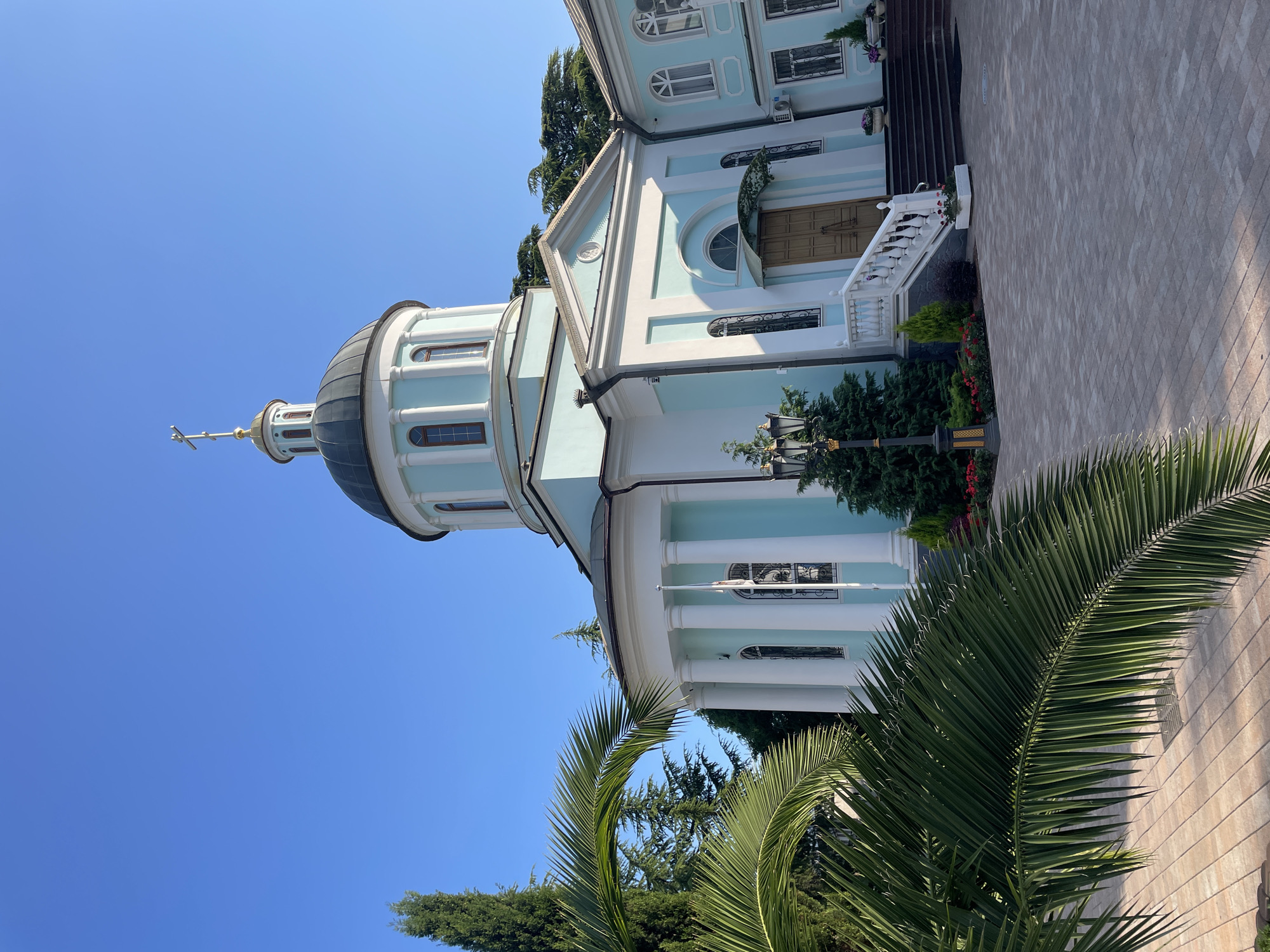 The image shows a light blue and white domed building, possibly a church or a historical structure, set against a clear, bright blue sky. The building features a prominent dome topped with a cross and several arched windows. A set of white steps with a railing leads up to a wooden door. The surrounding grounds are landscaped with lush green trees, including palm fronds in the foreground, and flowering bushes. The paving stones on the right side of the image suggest an outdoor pathway. A lamppost with an ornate design is visible near the building. The lighting indicates it is daytime, likely sunny, given the brightness and the clear sky. There is no visible text in the image. The scene appears peaceful and well-maintained, evoking a sense of tranquility. The orientation of the image is rotated.