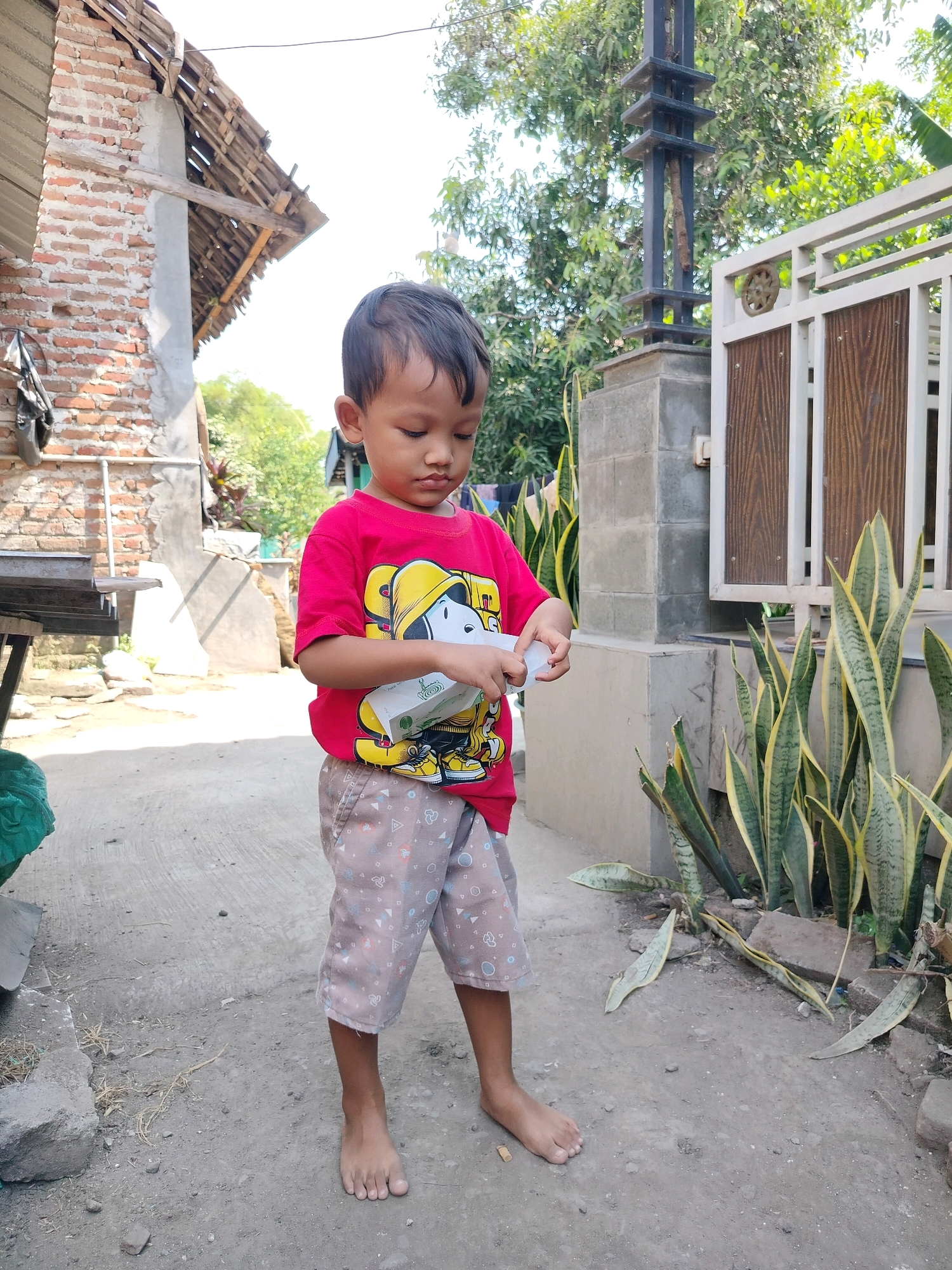 A young boy stands barefoot on a dirt path, looking down at a white object he holds in his hands. He wears a bright red t-shirt with a cartoon character graphic and light brown shorts with a patterned print. To his left is a brick building with a rustic wooden roof. To his right, a tall black metal pole stands next to a white gate with wooden inserts. Behind the gate, lush green trees and plants are visible. The environment appears to be an outdoor area in a residential setting, likely in Beloh, Indonesia, given the architectural style and vegetation.