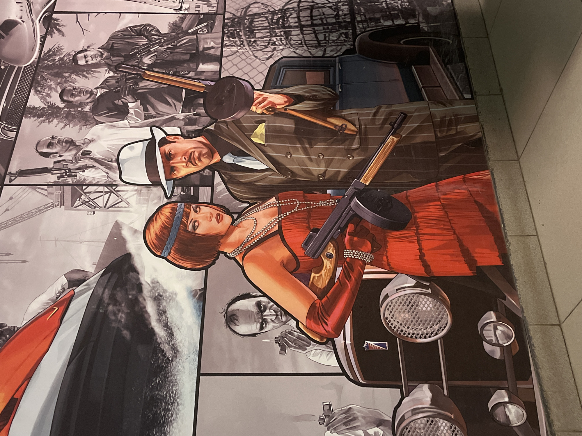 The image is a mural depicting characters from the Grand Theft Auto V video game. In the foreground, a woman in a red flapper dress and a man in a pinstripe suit are prominently featured. The woman holds a Tommy gun, and the man holds a similar weapon. Behind them, a vintage car is visible, along with other figures from the game, including men with firearms. The mural has a segmented composition, with different scenes and characters arranged in panels. The background suggests an urban or industrial setting with cranes and trees. The mural appears to be located in the city of Chaikovsky, Russia, likely as a decorative element on a building or in a public space. The artwork is stylized and illustrative, capturing the aesthetic of the video game.
