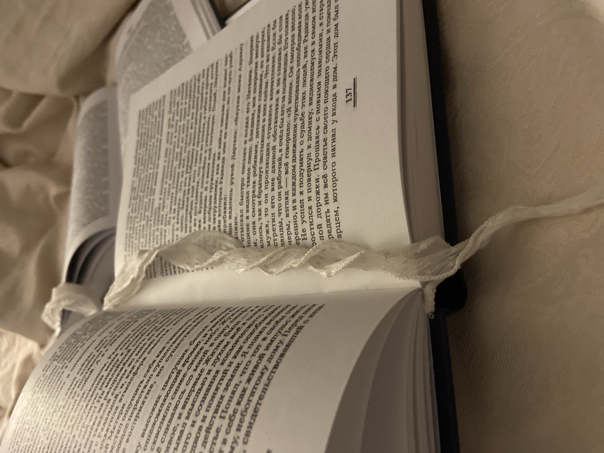 An open, dark-bound hardcover book with off-white pages featuring black Cyrillic text is centrally positioned. Page 137 is visible at the bottom of the right page. A white, textured fabric bookmark runs vertically along the book's spine. A second, plain white ribbon bookmark is partially visible further into the book, in the upper right. The book rests on a light beige, textured textile surface. Additional blurred open books are in the background. The scene is illuminated by warm, indirect light, indicative of an indoor setting.