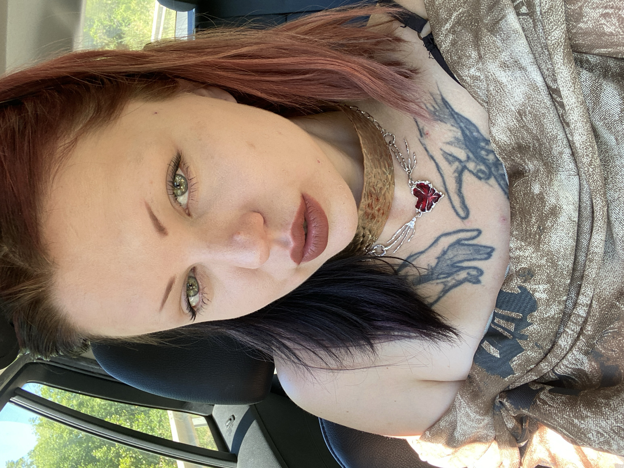 A woman with green eyes and two-toned hair (red on top, black underneath) is looking at the camera. She has tattoos on her chest and is wearing two necklaces. She is inside a car; the window shows green trees and a bright sky. The location, Kruče-Kërruçi, Montenegro, is not directly visible.