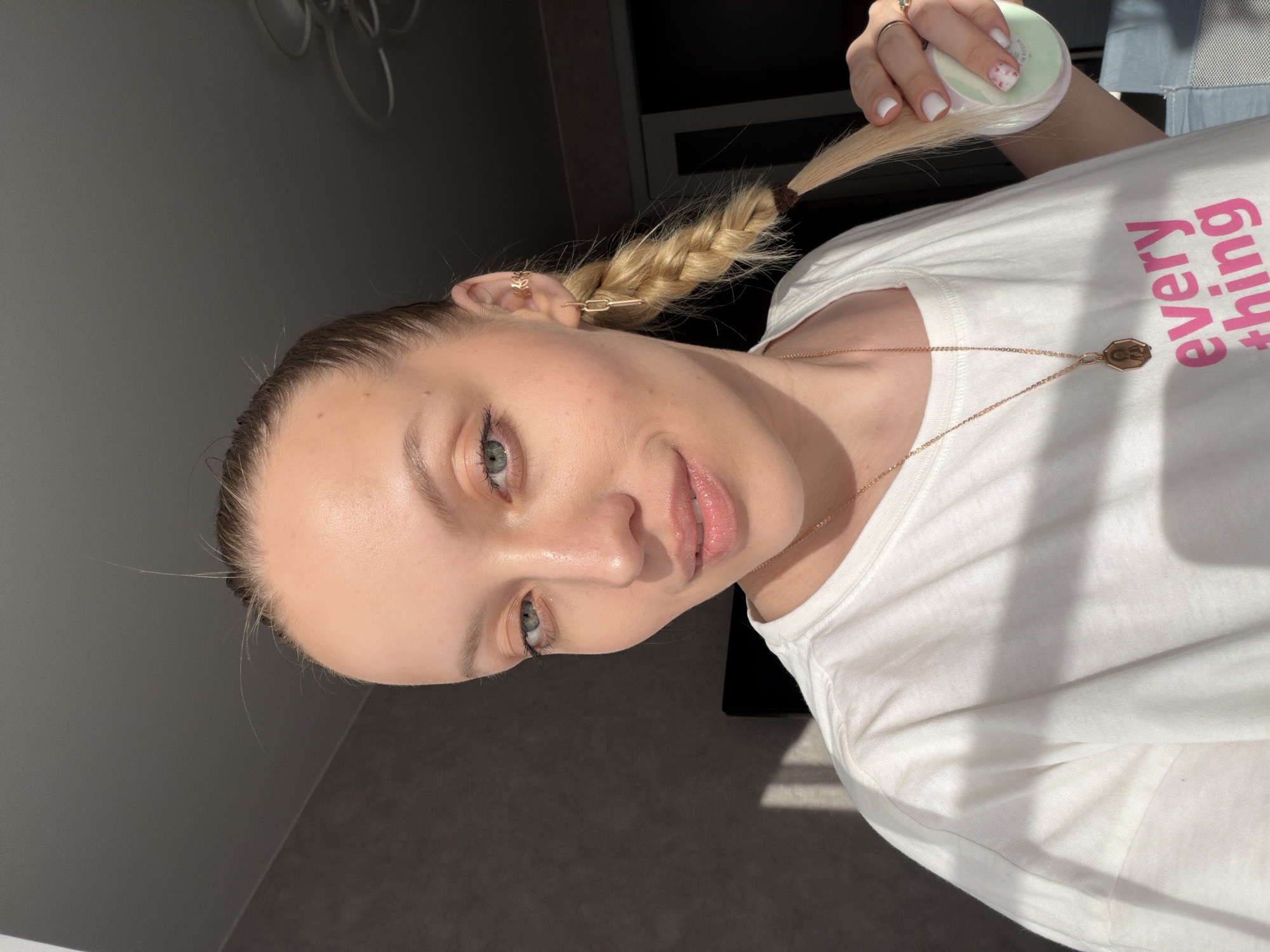This image presents a head-and-shoulders portrait of a light-skinned female individual, located indoors in Reshetnikovo, Russia. She has blue-green eyes and light blonde hair, pulled back from her face, with a single braid extending over her right shoulder. Fine, short hairs are visible around her forehead. She wears a white short-sleeved t-shirt with a partial pink inscription "every thing" on the front. Her accessories include a gold-colored chain necklace with an elongated, hexagonal pendant featuring a figure, a gold-toned linked drop earring in her right ear, and a gold-toned decorative ear cuff on the upper cartilage of her right ear. Her left hand, with white polished fingernails that include a pink speckled pattern on the ring finger, is shown holding a circular, light green compact object while also grasping the end of her braid. She is looking directly towards the camera with a subtle upturn of the lips. The immediate background comprises a dark gray wall, marked by a rectangular patch of direct sunlight on the left. The upper right background reveals a white ceiling and a multi-armed light fixture. A dark rectangular object and a light blue textile item are partially visible in the distant background.