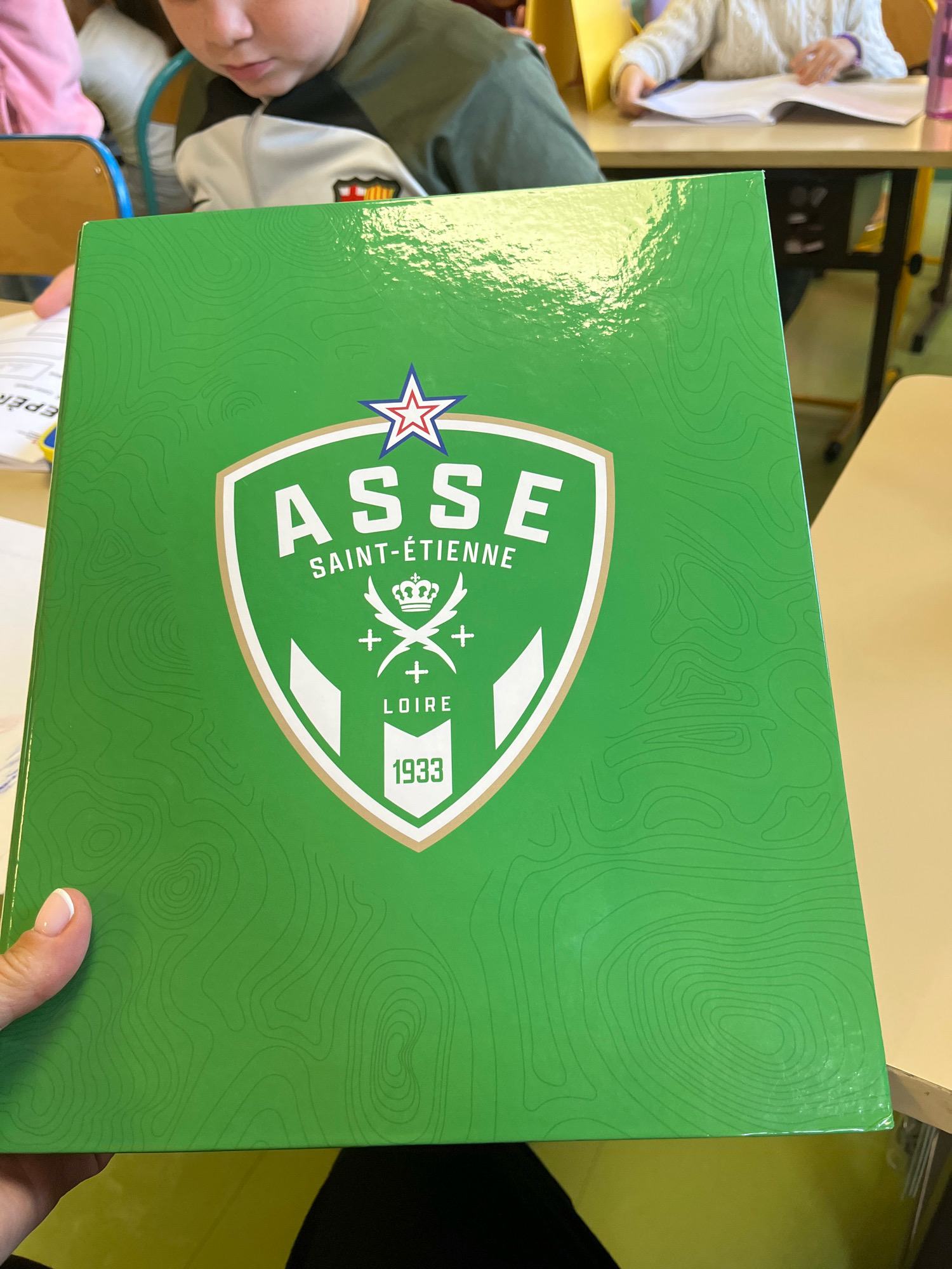 The image shows a close-up of a green binder with a crest featuring "ASSE SAINT-ÉTIENNE LOIRE 1933" in white text. The crest is bordered in gold and white. The binder's surface has a pattern of topographical lines. In the background, out of focus, are children sitting at desks in what appears to be a classroom setting. One child in the foreground is partially visible, wearing a dark green and white jacket. Another child in the background is wearing a white knitted sweater and is seen looking at a book. The overall scene suggests a school environment. The presence of a binder with the ASSE logo implies this might be in a school in or around Saint-Étienne, a city in France. La Fouillouse is a commune located in the Loire department, in the Auvergne-Rhône-Alpes region of central France, which is consistent with the area associated with ASSE.