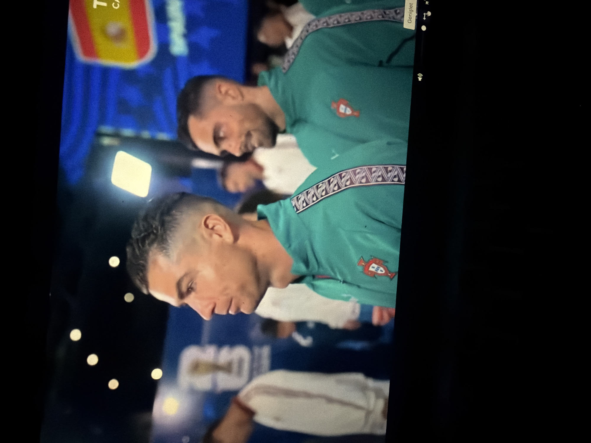 The image captures a close-up of two men wearing matching teal athletic jackets with a patterned trim. The man on the left, in the foreground, is identified as Cristiano Ronaldo due to his distinctive facial features and haircut. He is looking to his left, with a serious expression. He is wearing a teal jacket with a Portuguese crest on the left chest. The man to his right, partially visible, is also wearing a similar jacket and looking towards his left with a slight smile. The background is blurred and appears to be an indoor setting with blue and yellow banners, one of which displays the Spanish flag. There are also out-of-focus figures in the background. The lighting suggests an artificial environment, possibly a stadium or an event venue. The image is a still frame from a video, as indicated by the presence of video playback controls at the bottom right, including a "Genişlet" button, which is Turkish for "Expand." The image does not contain any information indicating a location in Nikopol, Bulgaria.