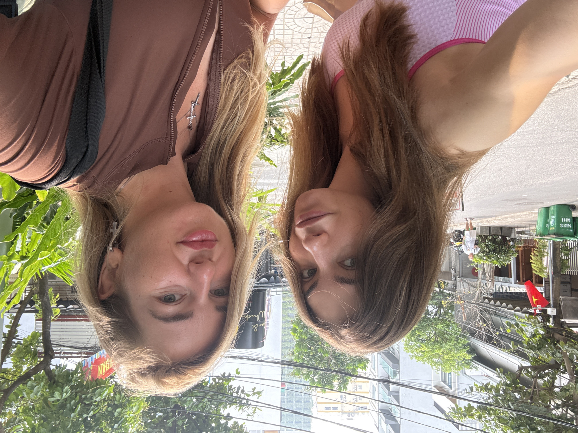 The image, rotated 90 degrees counter-clockwise, captures a close-up selfie of two women posing outdoors on a sunny day in Đà Nẵng, Vietnam. Both women gaze directly at the camera with neutral to slightly smiling expressions.
On the left, a woman with blonde hair wears a brown, long-sleeved zipped top. A black strap, possibly from a bag, crosses her shoulder. She sports a silver necklace featuring a sword-like pendant adorned with small crystals and a small, dark teardrop-shaped charm.
To her right, another woman with brown hair is visible, wearing a pink and white striped top. Her right arm is partially visible, suggesting she might be holding the camera or being photographed alongside her companion.
The background reveals a bustling urban street scene typical of Vietnam. Lush green trees and foliage are abundant, providing shade. Numerous power lines crisscross the sky, and several multi-story buildings are visible in the distance. The bright sunlight indicates it is daytime, likely midday or early afternoon.
Notable details confirming the location include a red flag with a yellow star, the national flag of Vietnam, visible on the right side of the street. Nearby, a green trash bin has white text on it, partially legible as "BINH" and "MISS SUI". Another sign in the background, glimpsed in a crop, displays "Wine & Whisky," and a red sign shows the partial word "KHAN," suggesting a local establishment. A person in a white t-shirt and shorts can be seen walking in the distance, further emphasizing the warm climate.