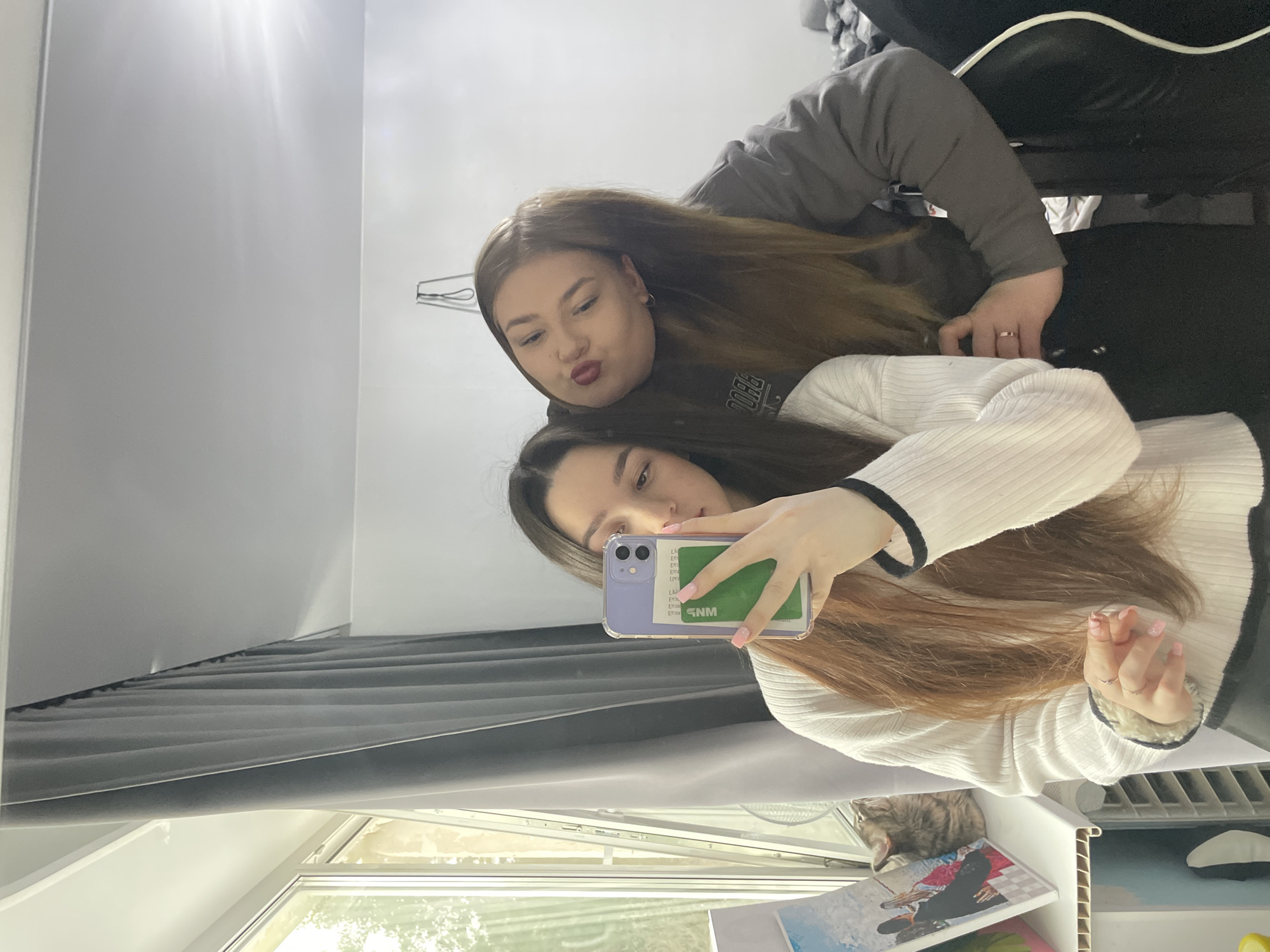 Two young women are taking a selfie. The woman in the foreground is holding a cell phone with a green rectangle covering the screen. She is wearing a white sweater with black trim. The woman in the background is making a kissing face and wearing a grey hooded sweatshirt. The location appears to be a residential room with a window looking outside, potentially in Chaikovsky, Russia. A cat is partially visible near the bottom of the frame.