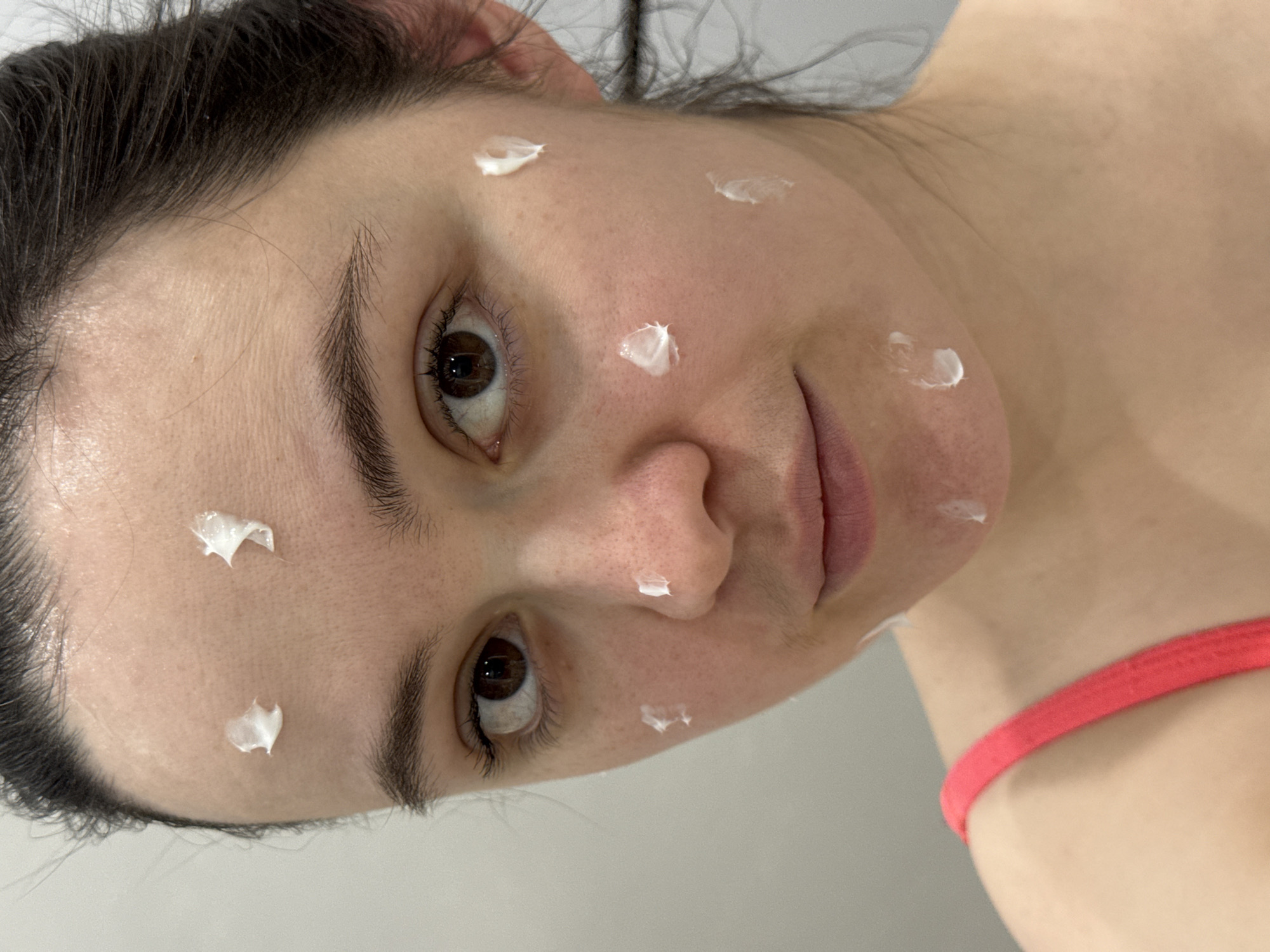 A close-up, slightly rotated shot shows a woman's face with dabs of white cream applied to her forehead, nose, and cheeks. She has dark hair pulled back, brown eyes, and a light complexion. A red strap of clothing is visible on her shoulder. There is no information to determine that this image was taken in the city of Izhevsk, Russia, therefore I can not use that in my response.