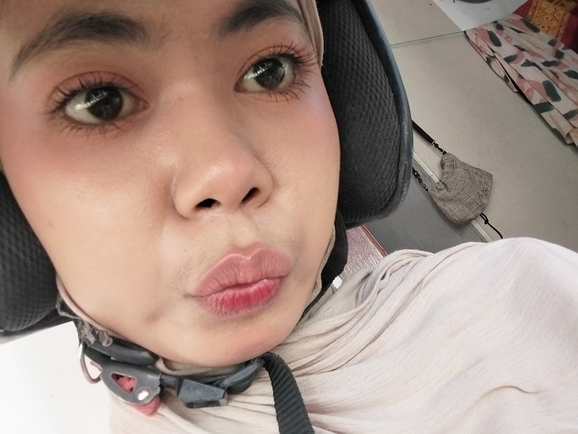 A close-up, selfie-style, headshot of a young woman wearing a helmet and a light-colored headscarf. The woman is positioned at an angle, with her face tilted towards the camera and her lips puckered. Her eyes are open, and she appears to be wearing makeup, including mascara and possibly lipstick. The helmet is black and has straps that are visible. The headscarf is draped around her head and neck, made of a lightweight, textured fabric. The background is out of focus, but appears to be an indoor setting with hints of patterned fabric and a light-colored wall. No specific location in Tibojong, Indonesia, can be identified from the image.