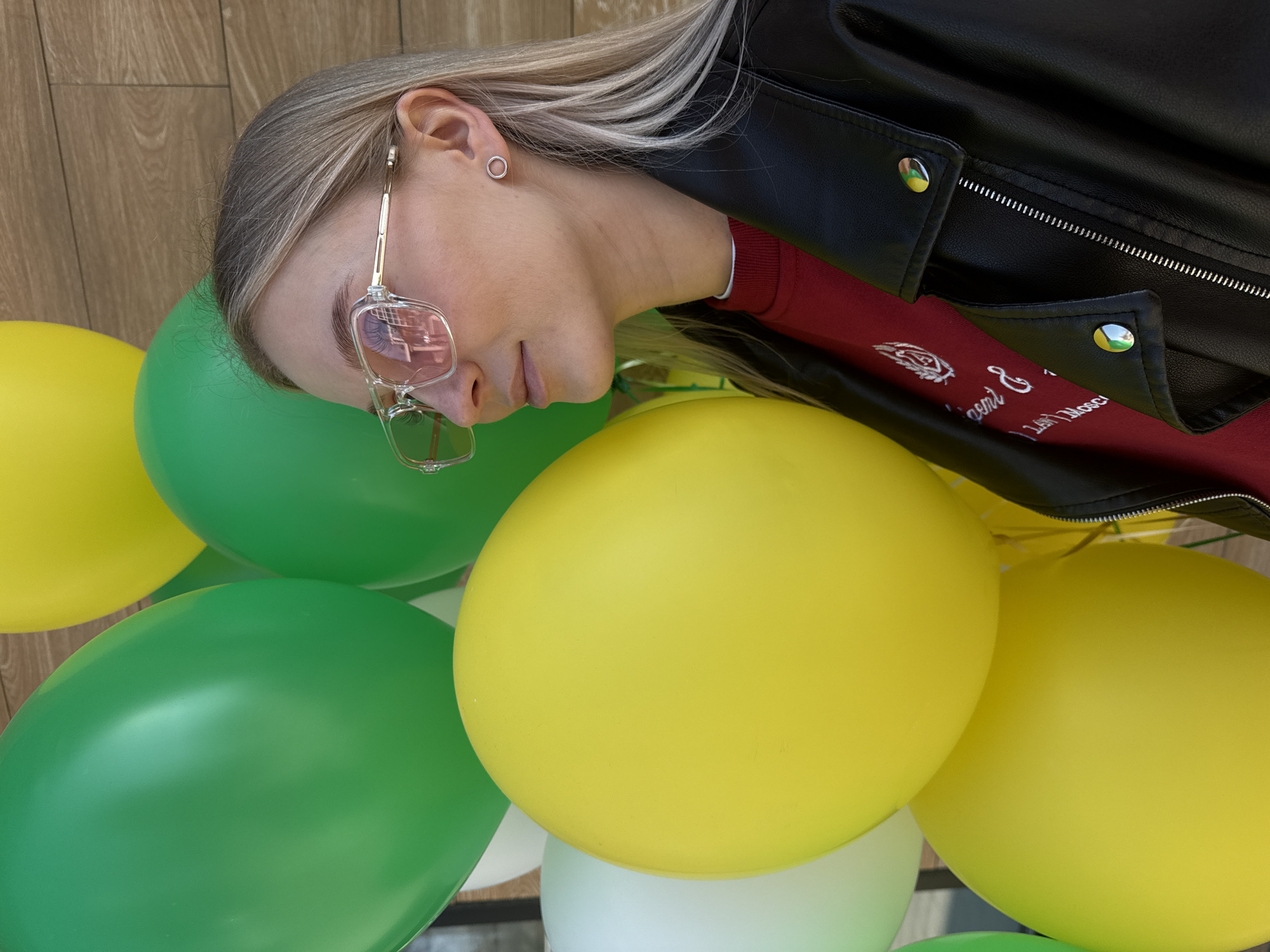 A young woman is holding a cluster of yellow and green balloons. She is wearing a black leather jacket over a red sweater and eyeglasses. The location appears to be outdoors, possibly in Izhevsk, Russia, although there are no explicit markers to verify the location.