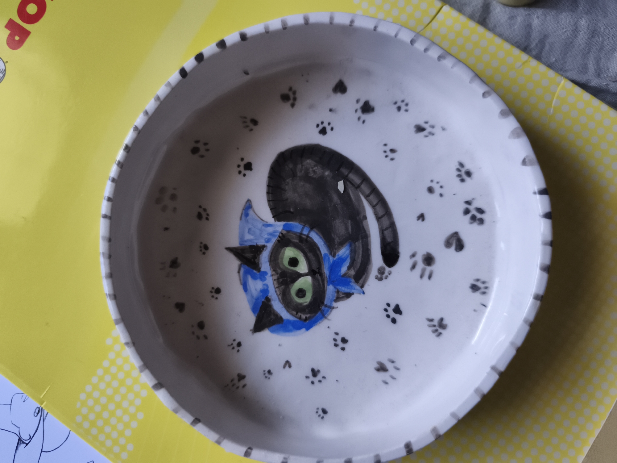 A white ceramic bowl decorated with paw prints and a cartoon cat is placed on a yellow surface with a polka dot pattern. The cat is blue with green eyes and black markings. The bowl has a black and gray striped rim. The yellow surface appears to be a placemat or a mat. The overall scene is viewed from directly above. The image does not depict the city of Saint-Denis, France.