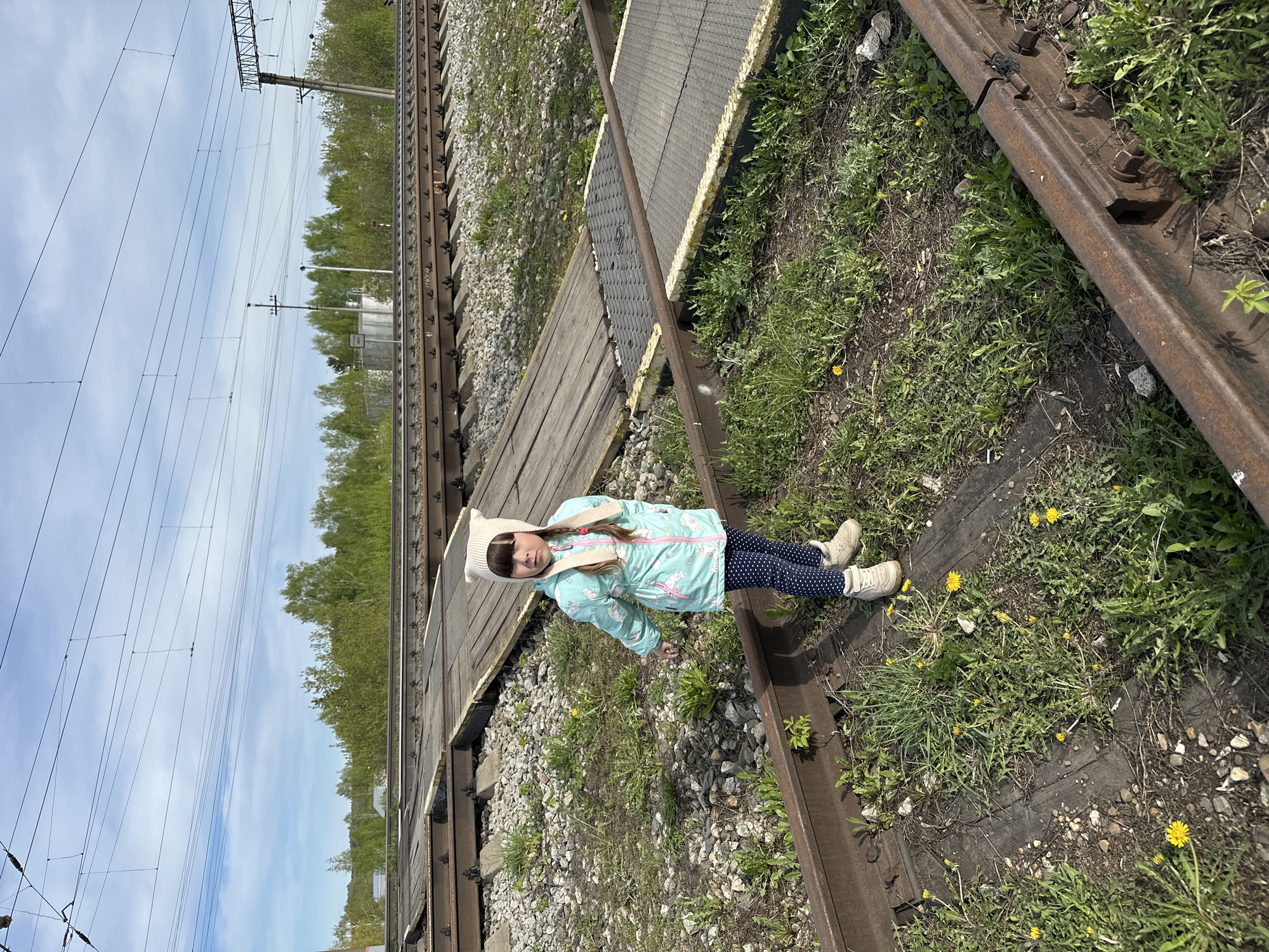 A young girl stands on a railway track in Кечёво, Russia. She is wearing a light blue jacket with a unicorn pattern, dark blue leggings with white polka dots, and light beige shoes. A white hat with animal ears is on her head. The railway track is overgrown with grass and small yellow flowers. A wooden walkway with a grey, textured section runs across the tracks. Railway lines and power lines are visible in the background. The sky is partly cloudy.