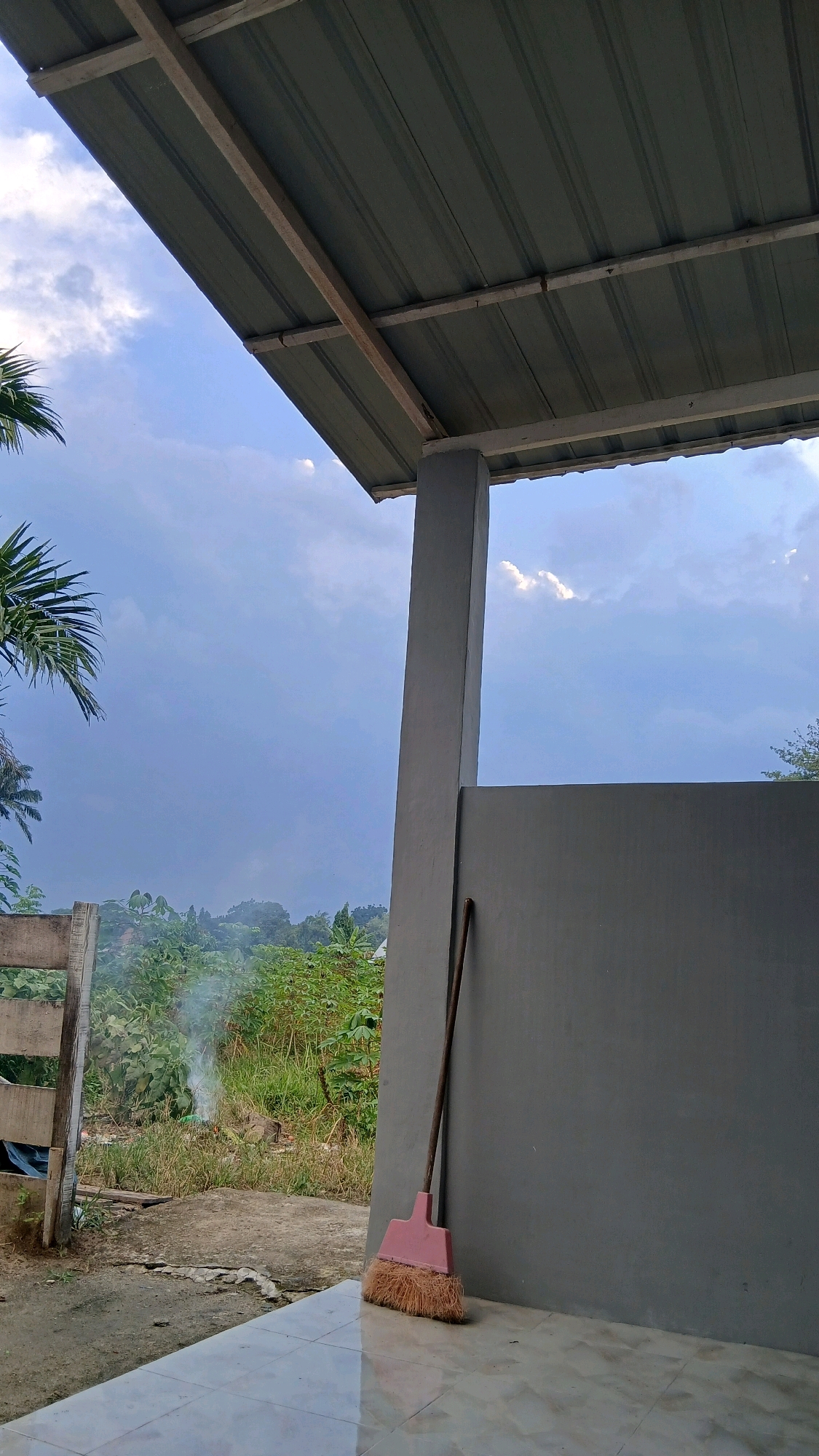 The image depicts an outdoor scene under a cloudy sky, likely in Hamparan Perak, Indonesia. In the foreground, a pink broom with a wooden handle leans against a grey wall. The ground is tiled and reflects the light. Beyond the wall, a wooden fence is visible, and behind that, lush green vegetation and trees stretch into the distance. A plume of smoke rises from the vegetation, suggesting an outdoor fire or burning. The sky is a mixture of blue and grey clouds, with some lighter patches indicating breaks in the cloud cover. The overall impression is of a rural or semi-rural setting.