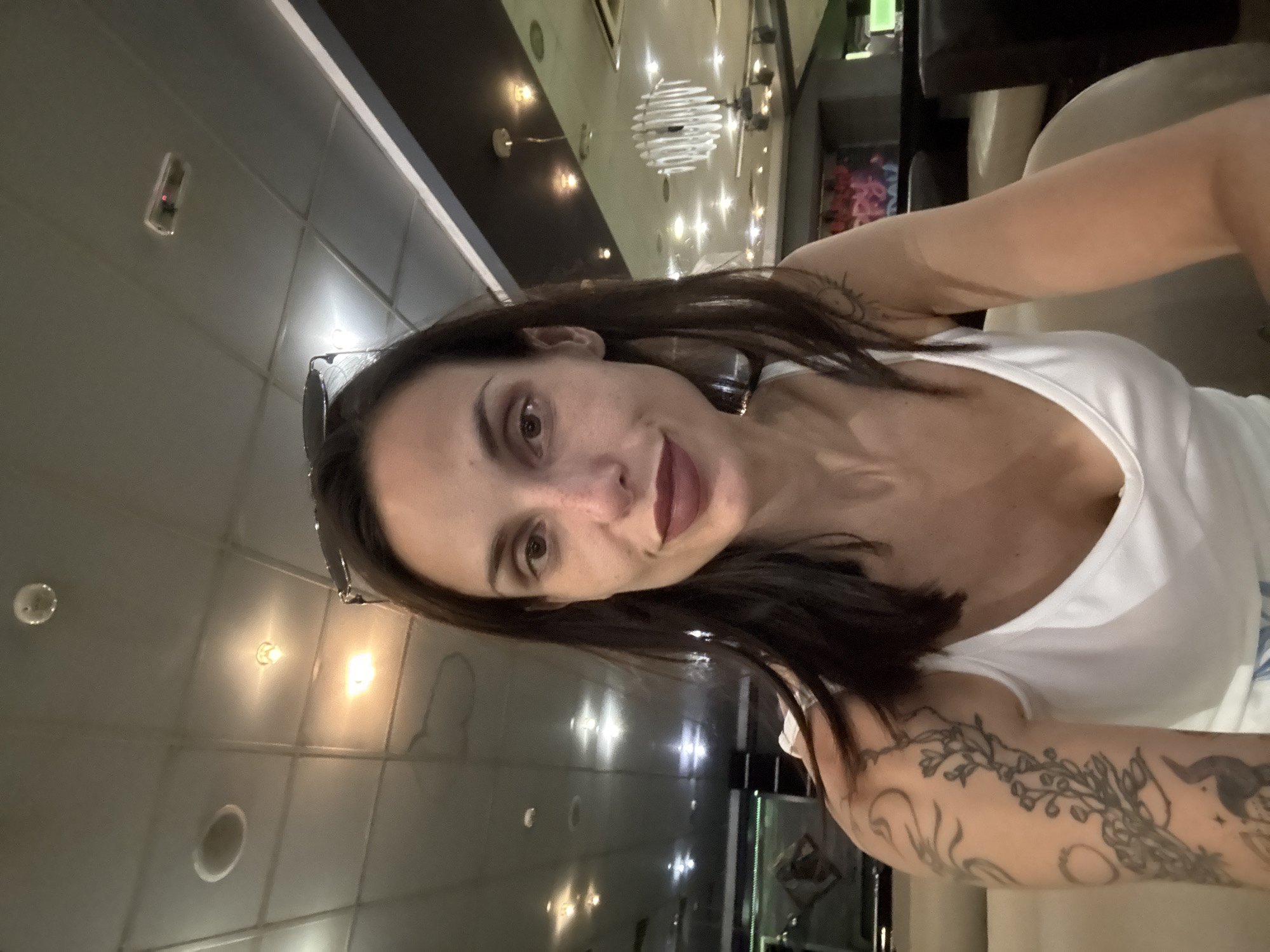 A woman with dark hair and dark lipstick is taking a selfie in an indoor setting. She is wearing a white tank top and has tattoos on her left arm. The background appears to be a lounge or restaurant with a tiled ceiling, recessed lighting, and a decorative chandelier. In the distance, there is a wall with graffiti and seating areas. The woman is seated and looking directly at the camera. The specific location in the city of Pleven, Bulgaria is not discernible from the image.