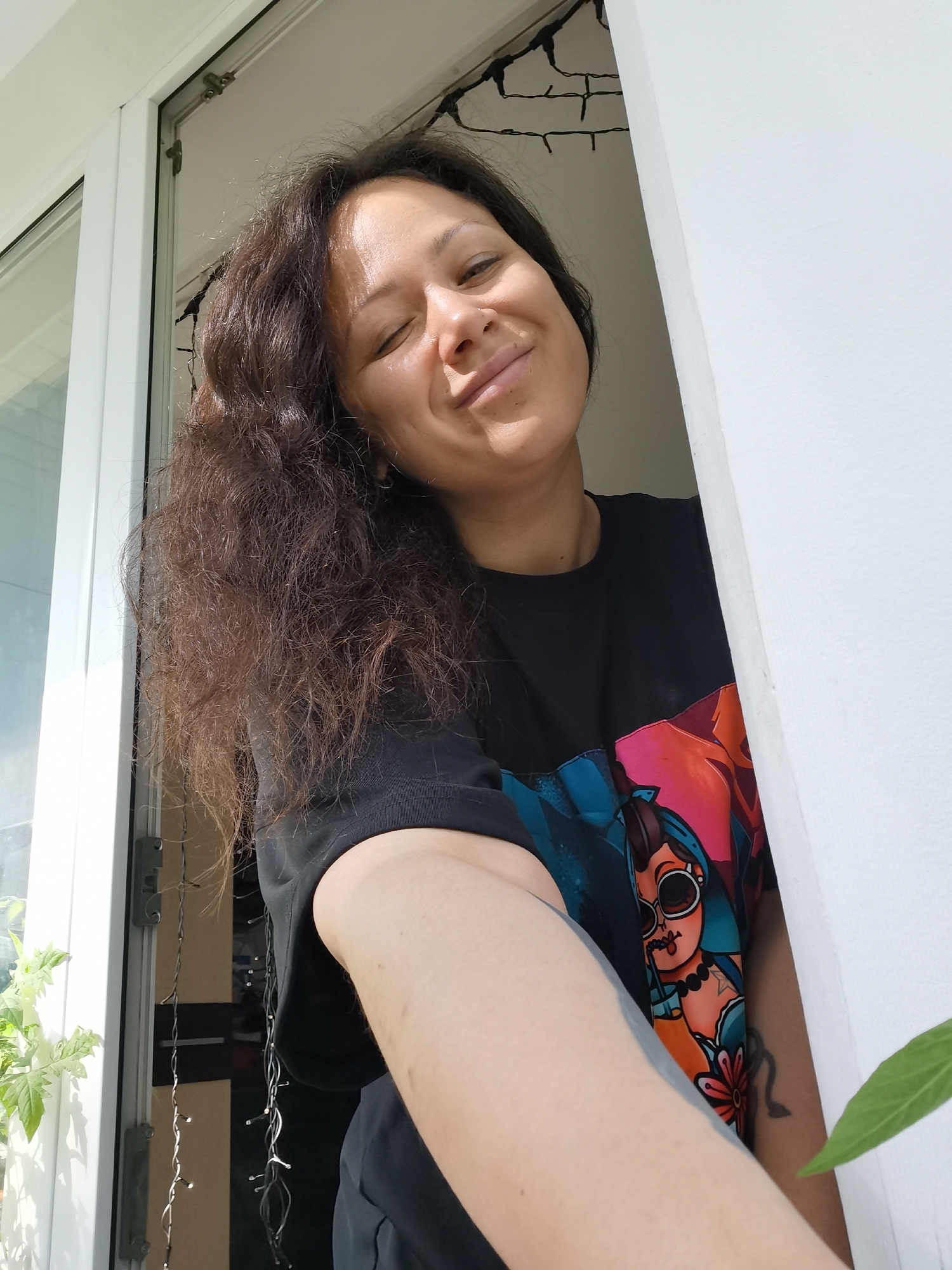 A woman with long, dark, wavy hair is smiling and looking slightly downward. She has a nose ring. She is wearing a black t-shirt with a colorful graphic print featuring a cartoon woman with sunglasses, drinking from a straw. The woman is standing near a white window frame. A string of lights hangs near the window. Green plants are visible outside the window. The location could be Chaikovsky, Russia.