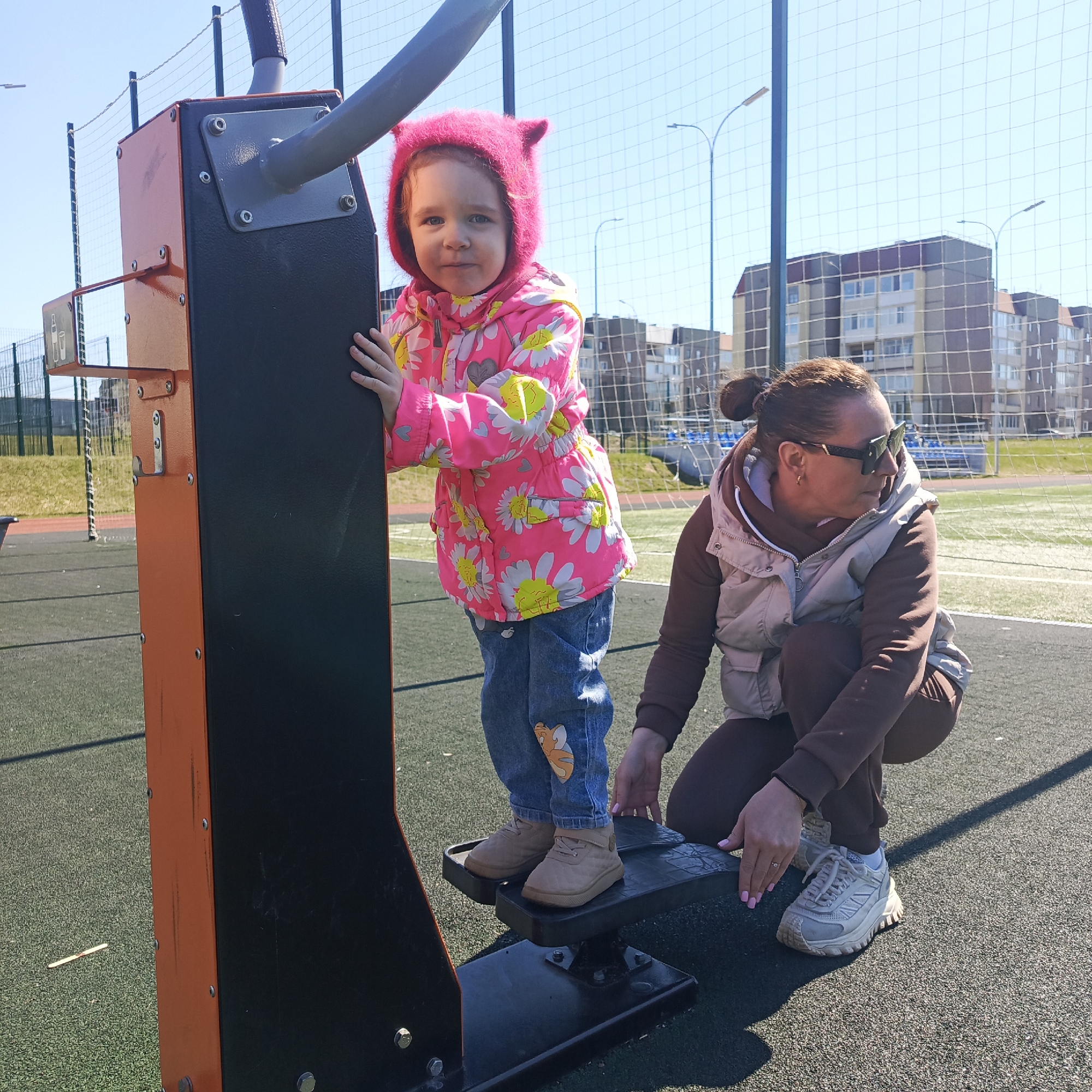 A young child in a pink floral jacket and jeans stands on a piece of playground equipment, gripping a black metal bar. An adult woman wearing sunglasses, a brown sweatsuit, and a beige vest crouches next to the equipment, seemingly helping the child. The equipment has orange sides and a black central part. In the background, a green athletic field and apartment buildings can be seen through a chain-link fence. The location is Марковский, Russia.