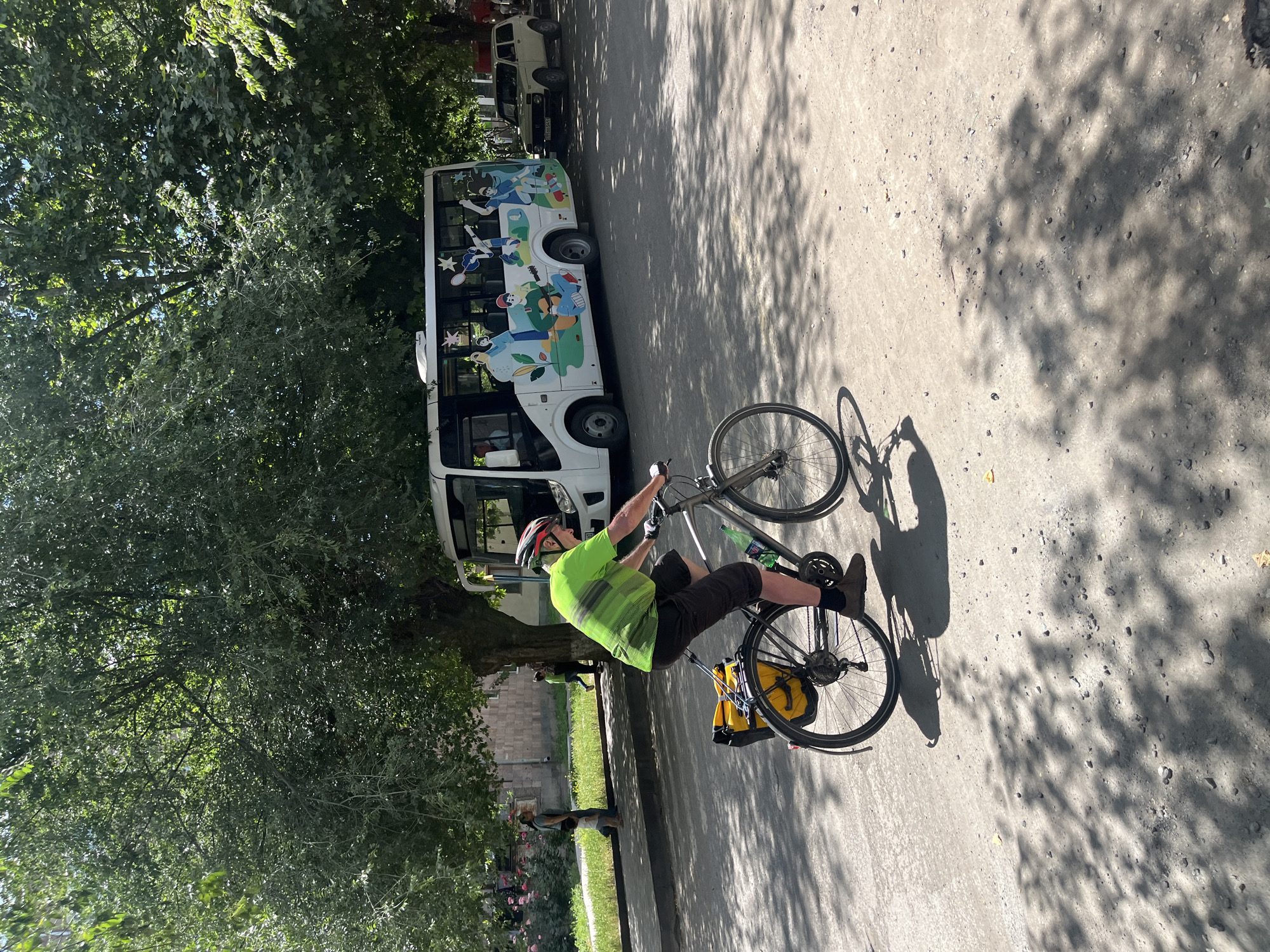 A man wearing a helmet and a lime green shirt with dark shorts is riding a bicycle. He is positioned in the center of the frame, facing away from the viewer. The bicycle has two yellow bags attached to the rear rack. The scene appears to be outdoors on a paved surface, possibly a street or pathway, with dappled sunlight casting shadows from trees. To the left of the man and bicycle is a white bus decorated with colorful cartoon characters engaged in various activities. The bus has tinted windows and appears to be parked or moving slowly. Further left, there are dense green trees providing shade. Two other people are visible in the distance, also appearing to be on bikes, and some pink flowers are present in the foreground. The lighting suggests it is daytime, likely a sunny day. The overall impression is one of a casual outdoor activity, possibly a scenic route or a park setting. No visible text is legible on the bus or elsewhere in the image.
