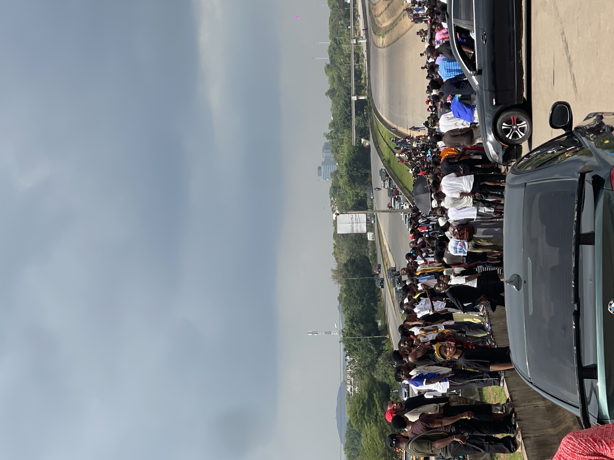 A large crowd of people are gathered on the side of a multi-lane highway in Abuja, Nigeria. The sky is overcast with dark gray clouds. In the background, a telecommunications tower stands tall, and further in the distance, several modern buildings can be seen amongst lush green trees. The highway has multiple lanes and appears to be elevated in some sections. Cars are visible on the road, some driving and others parked. Many of the people in the crowd are young men, dressed in casual clothing, standing along a concrete barrier. Some individuals appear to be holding umbrellas. Two cars are in the foreground, one black and one dark green, with a portion of their rear ends visible. The overall scene suggests a public gathering or event on a major road in Abuja.