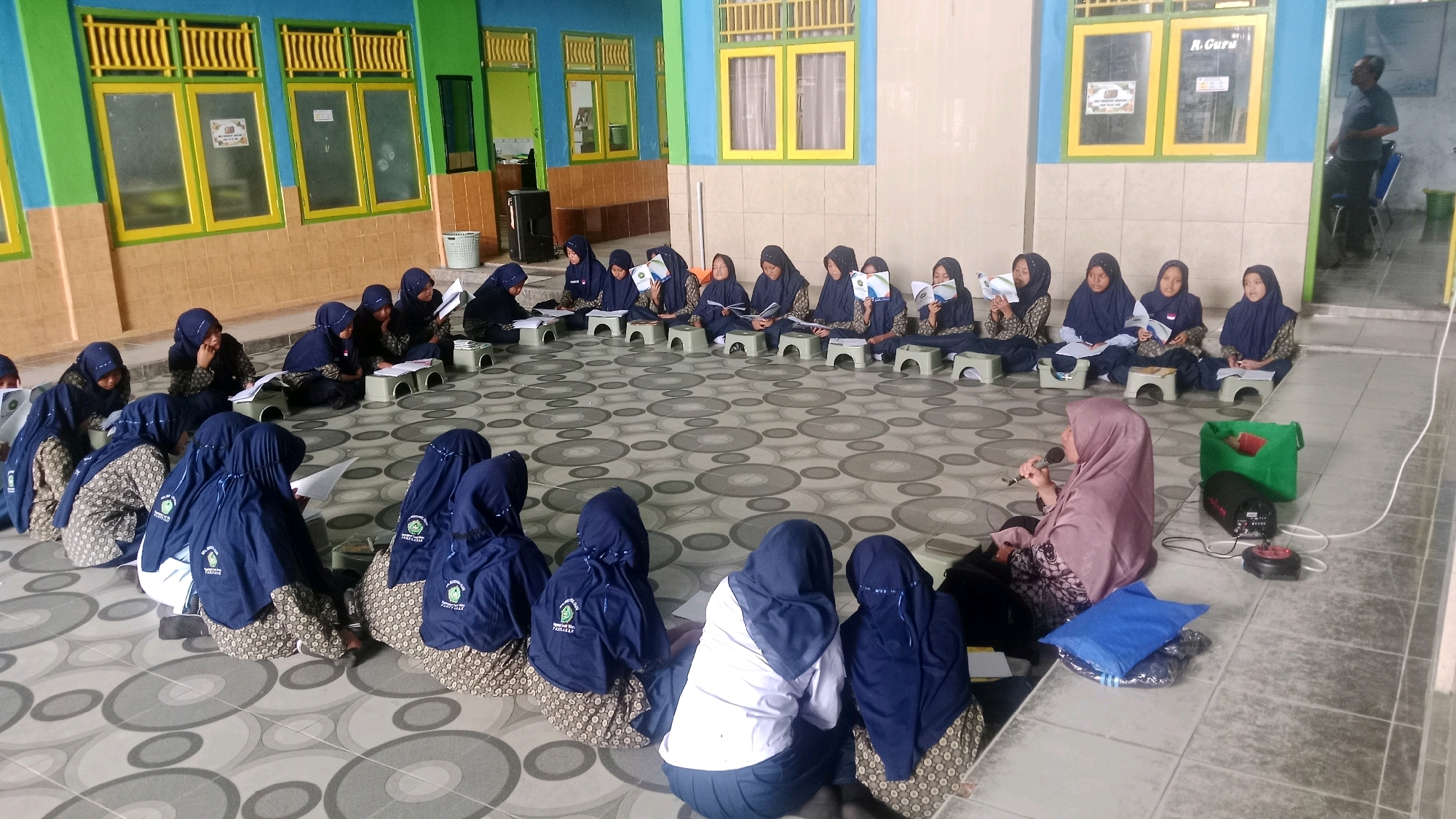 The image depicts an outdoor scene in Waru, Indonesia, likely at an educational institution, possibly a madrasah or school. A group of young students, predominantly female, are seated in a circular formation on a tiled floor. They are wearing dark blue headscarves (hijabs) and patterned skirts or sarongs. Many of the students are holding books or papers and appear to be engaged in a learning activity, possibly reading or listening.
In the foreground, a woman wearing a light purple hijab and a patterned dress is seated, holding a microphone and speaking, suggesting she might be a teacher or instructor leading the session. Several electronic devices, including what appears to be a speaker and a microphone system, are placed near her. A green bag and blue cushions are also visible on the floor.
The background shows a building with brightly colored walls – yellow and green – and windows with yellow frames. The overall setting appears to be an open-air or semi-enclosed courtyard area adjacent to the building. The tiled floor features a geometric pattern of grey circles. The presence of the students in uniform, the teacher with a microphone, and the educational materials strongly indicate a school or religious education setting.