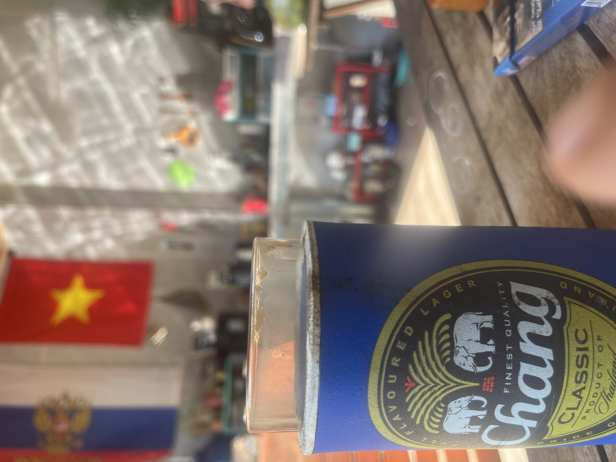 A Chang beer can cooler is prominently displayed in the foreground of the image, resting on a wooden table. The can cooler is blue with the Chang logo, featuring two elephants and text that reads "FLAVOURED LAGER," "Chang," "FINEST QUALITY," "CLASSIC," and "PRODUCT OF Thailand." A clear plastic lid sits atop the can cooler. In the background, a blurred scene shows two flags hanging, one red with a yellow star (likely the Vietnamese flag) and another with blue and white stripes and a crest. Various objects, possibly kitchen items or shelves, are visible in the further background, with sunlight casting patterns on a wall. The overall setting appears to be an outdoor or semi-outdoor area, consistent with a location in Phan Thiet Ward, Vietnam.
