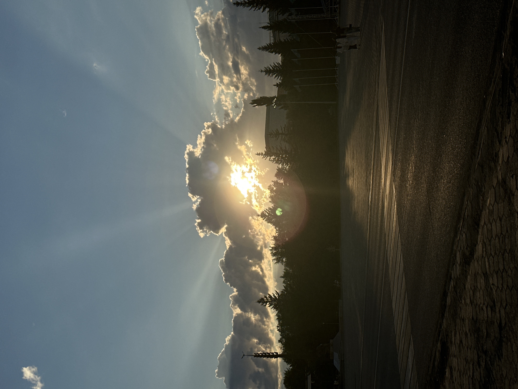 The image depicts a silhouetted landscape at sunset in Kokshetau, Kazakhstan. The sun is setting behind a line of trees and clouds, casting dramatic rays of light across a bright blue sky. In the foreground, a paved area with a crosswalk suggests a public space, possibly a plaza or a street. A few figures are visible in the distance, walking across the paved area. The overall atmosphere is one of tranquility and natural beauty, enhanced by the warm glow of the setting sun.