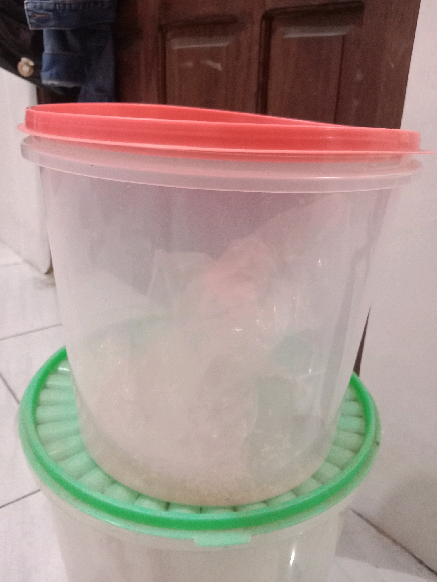A close-up, eye-level shot showcases two translucent plastic containers stacked vertically, both with clear plastic bodies and different colored lids. The top container has a bright pink lid and contains a powdery white substance, possibly sugar or flour, filling about a third of its volume. The lower container is partially obscured but appears to have a light green lid with a ridged pattern. The containers rest on a tiled floor, and the background reveals a dark brown wooden door and a hint of denim fabric, suggesting a domestic indoor setting. The image does not provide any information that would place it in Brangsong, Indonesia.