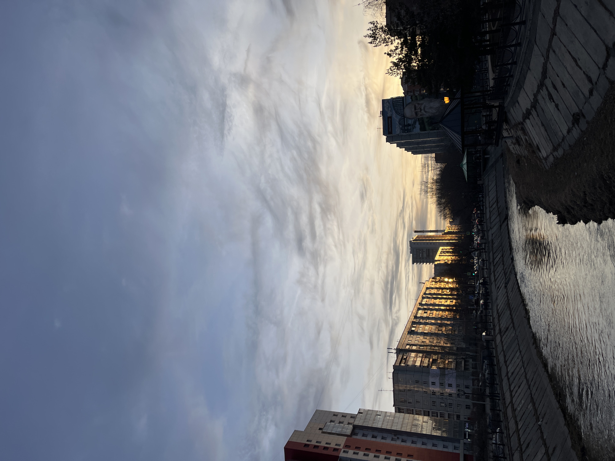 The image shows a view of the Yenisey River embankment in Krasnoyarsk, Russia, at dusk or dawn. The sky is filled with dramatic clouds, with light filtering through, creating a golden glow. Several buildings line the embankment, with their windows reflecting the sunlight. The river flows along a concrete embankment, and its surface reflects the buildings and the sky. Trees are visible along the riverfront.