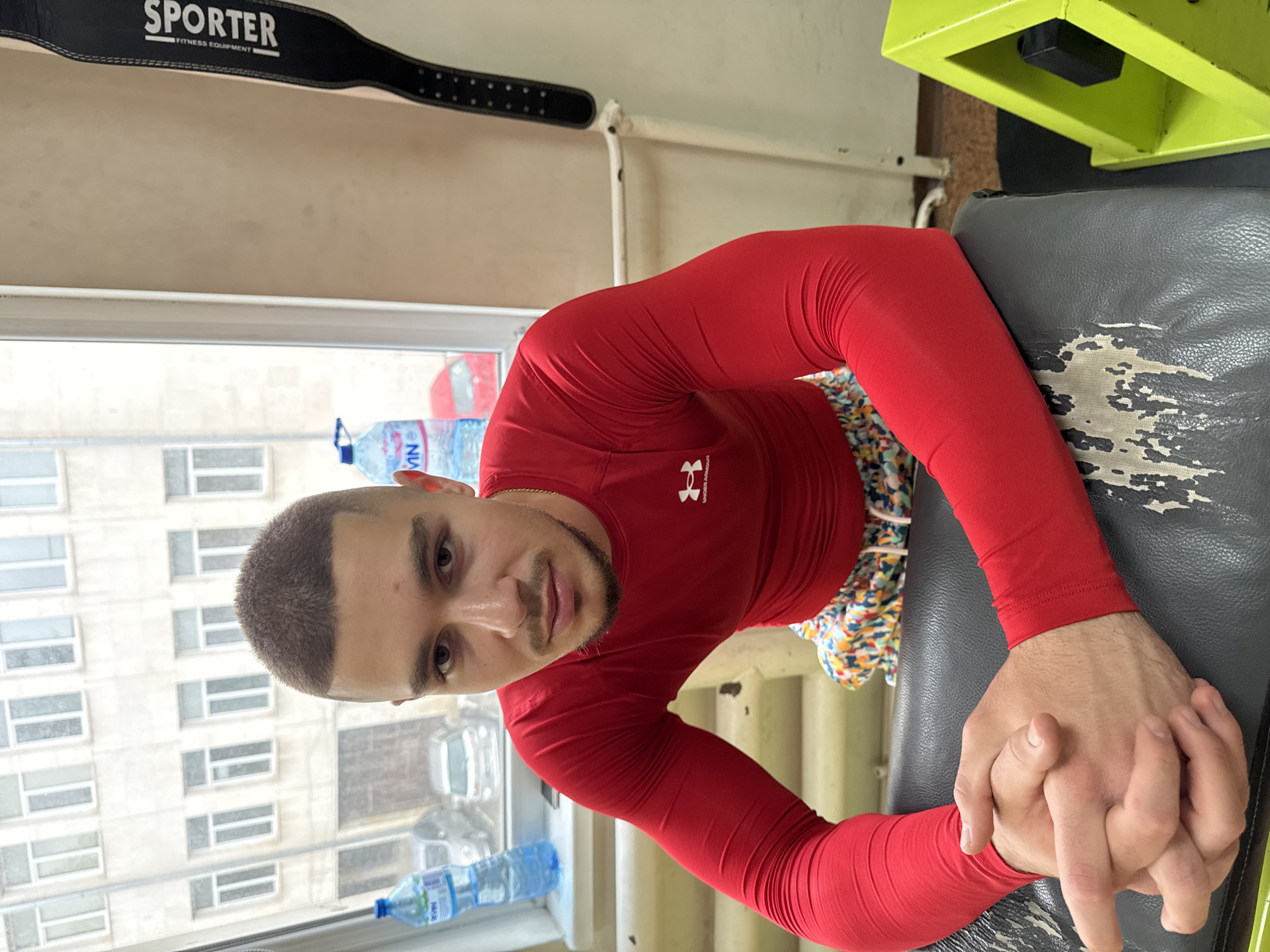 The image shows a man in a red Under Armour long-sleeved shirt and colorful shorts, sitting on a worn, gray cushioned seat, possibly part of a gym machine. He is clasping his hands together. He appears to be in a room with a large window overlooking a street with buildings. Bottles of water are visible on the windowsill. A black "SPORTER Fitness Equipment" belt is on a shelf. A partially visible green machine or structure is present near the seat.
The city of Pleven, Bulgaria, is not explicitly confirmed from the image alone, but it may be inferred from context outside of the visual information provided. No specific Pleven location can be determined from the image.