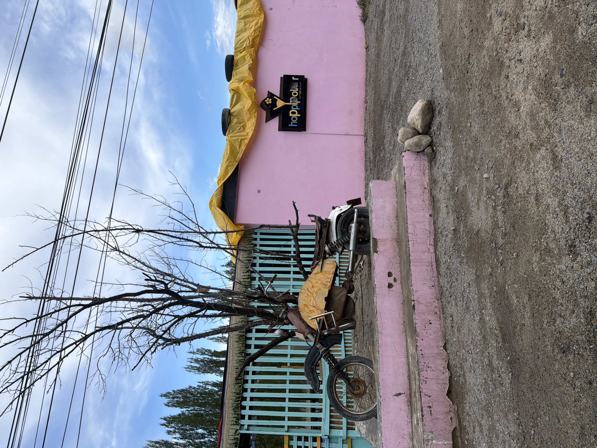 The image shows a pink building with a yellow tarp covering the roof. A sign on the building reads "hoppipola" with the tagline "EAT. DRINK. PLAY." A motorcycle, appearing to be a sculpture or art installation, is placed on a pink platform in front of the building. The motorcycle is weathered and has a yellow cover over its engine. A bare tree with no leaves stands behind the motorcycle. A light blue fence is visible to the left of the building, with tall trees in the background. The ground is covered in gravel and dirt. The scene appears to be outdoors, likely in a semi-urban or rural area, consistent with a location in Leh, India, where "Hoppipola" is a known entertainment venue. The sky is partly cloudy. There are no people visible in the image.