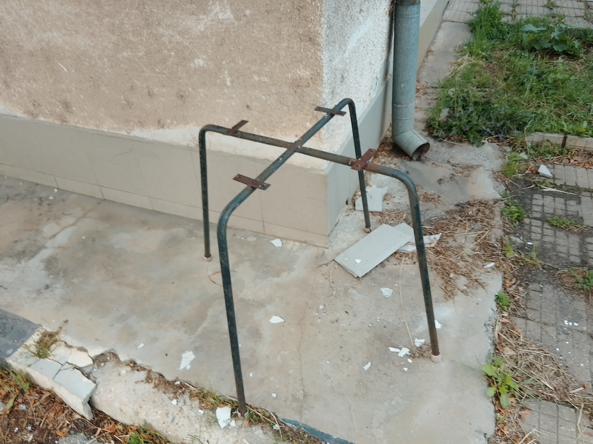The image captures an outdoor scene, primarily featuring a metallic frame placed next to a building wall. The main subject is a dark-colored metal frame, constructed from tubular metal bent into four legs that curve outwards at the top and bottom. Two flat metal crossbars connect the top sections of the legs, and there are additional rusty, flat metal pieces welded or attached to these crossbars, possibly as supports or attachment points. The frame appears sturdy but also somewhat weathered.
The frame rests on a concrete surface which shows cracks and discoloration. Adjacent to this concrete, a building wall rises. The lower part of the wall is clad with light-colored tiles, while the upper section has a rough, textured, light-brown or beige finish, showing signs of wear and age. To the right of the frame, a grey metal drainpipe runs vertically down the wall, bending outwards near the bottom.
Beyond the concrete and the drainpipe, the ground transitions to a mix of paved sections, covered with patches of grass, weeds, and dry foliage, indicating an unkempt area. Scattered white debris, possibly fragments of plaster or broken tiles, are visible on the concrete and among the vegetation. The lighting suggests daytime, but there are no strong shadows, indicating either an overcast sky or indirect light. No people are visible, and no specific activity is taking place. There is no visible text in the image. The overall setting appears to be a utilitarian or neglected outdoor space, possibly a side of a building or a back alley.
