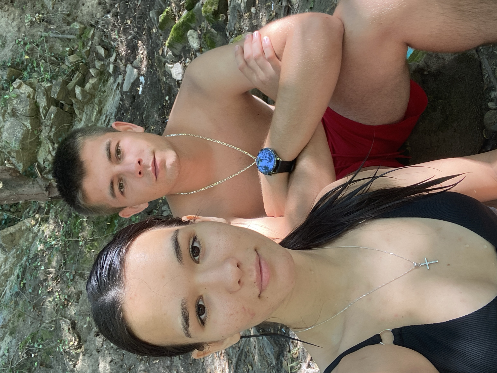 In Debnevo, Bulgaria, a young couple poses for a selfie outdoors, surrounded by rocks and foliage. The woman, in the foreground, wears a black bikini top and a silver cross necklace. Her dark hair is wet and pulled back. The man, behind her, is shirtless and wears red shorts and a silver chain necklace. He has a watch on his left wrist. They appear relaxed and are looking at the camera. The background suggests a natural, somewhat rugged environment with trees and moss-covered rocks.