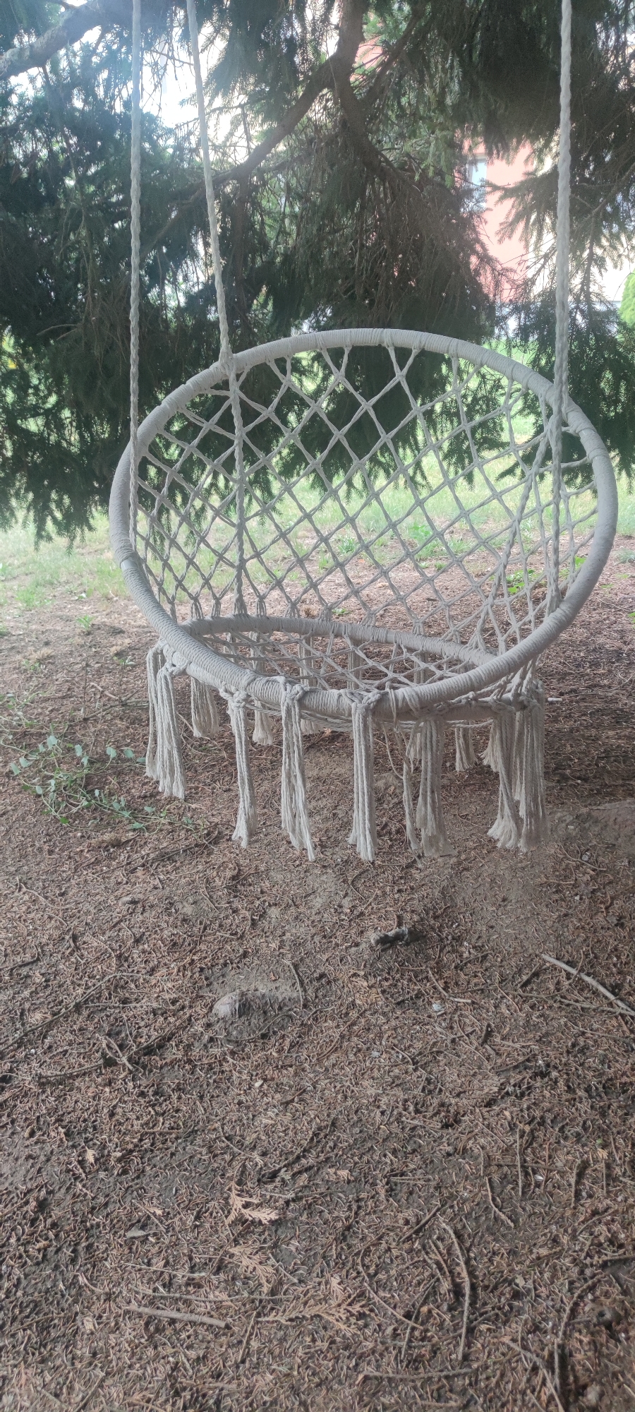 This media file shows a white macrame hammock chair hanging from the branches of a large tree. The chair is oval-shaped with a diamond-patterned weave and fringe tassels hanging from its bottom edge. Two thick ropes suspend the chair from the tree. The surrounding setting is outdoors, with dry, brown pine needles and small branches covering the ground beneath the chair. Lush green foliage from the tree forms a canopy overhead, and in the background, through the branches, a building with a reddish-pink facade and white window frames is partially visible. The lighting suggests it is daytime, possibly overcast or late afternoon, as there are no strong shadows. The scene appears to be a tranquil, natural environment, likely a backyard or park in Liebenburg, Germany, given the context. There is no text visible in the image.