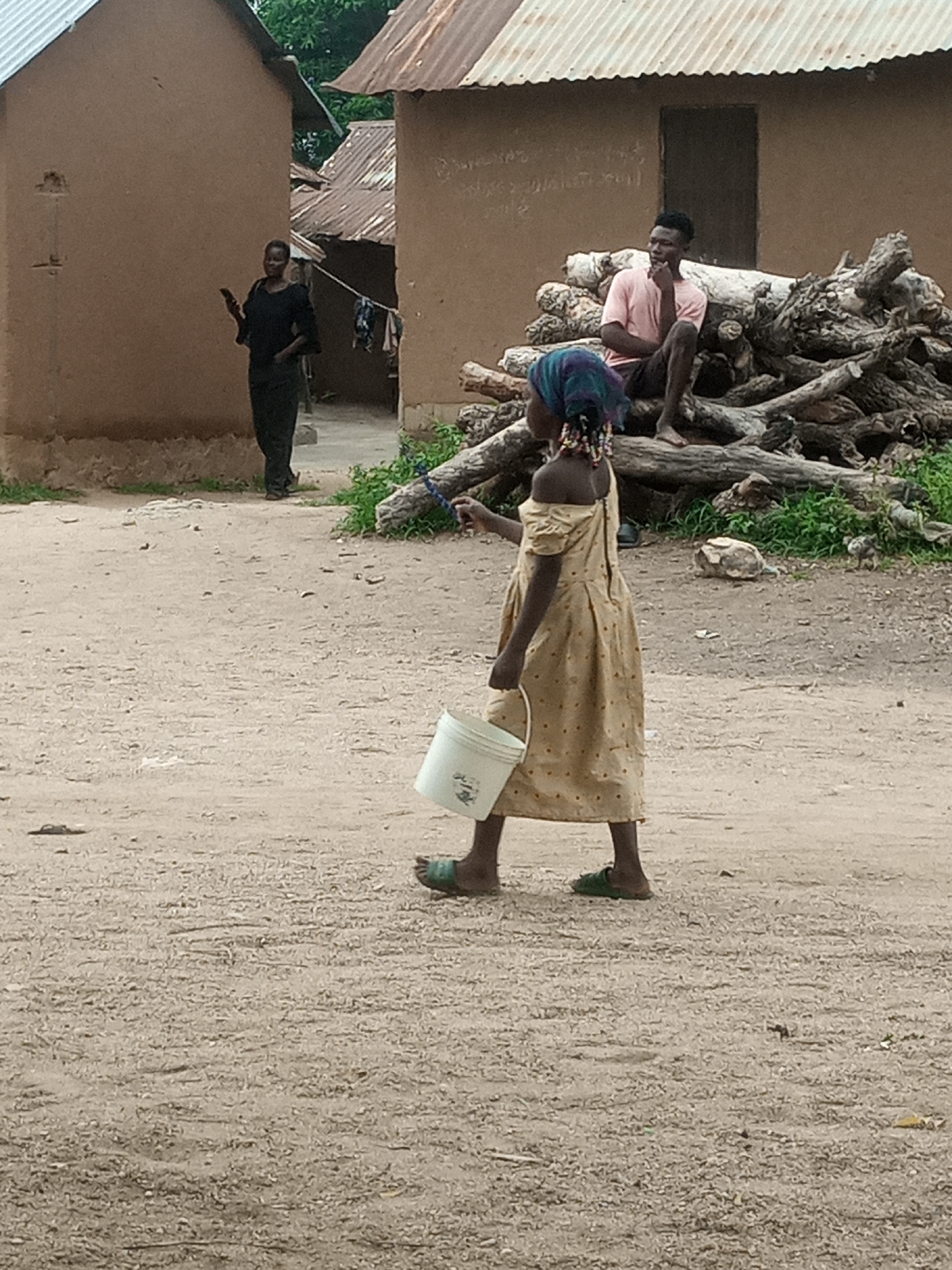 In an outdoor, rural setting, a young girl wearing a light-colored dress with red polka dots and a colorful headscarf walks from right to left, carrying a white bucket in her right hand. Her left arm is extended slightly forward. She wears green sandals on her feet. In the background, a man in a pink t-shirt and shorts sits on a pile of logs. Further to the left, a woman in a black dress stands with a mobile phone in her left hand. Two mud-brick buildings with corrugated metal roofs are visible behind the people. The ground is composed of dirt and small stones, with patches of green grass and vegetation.