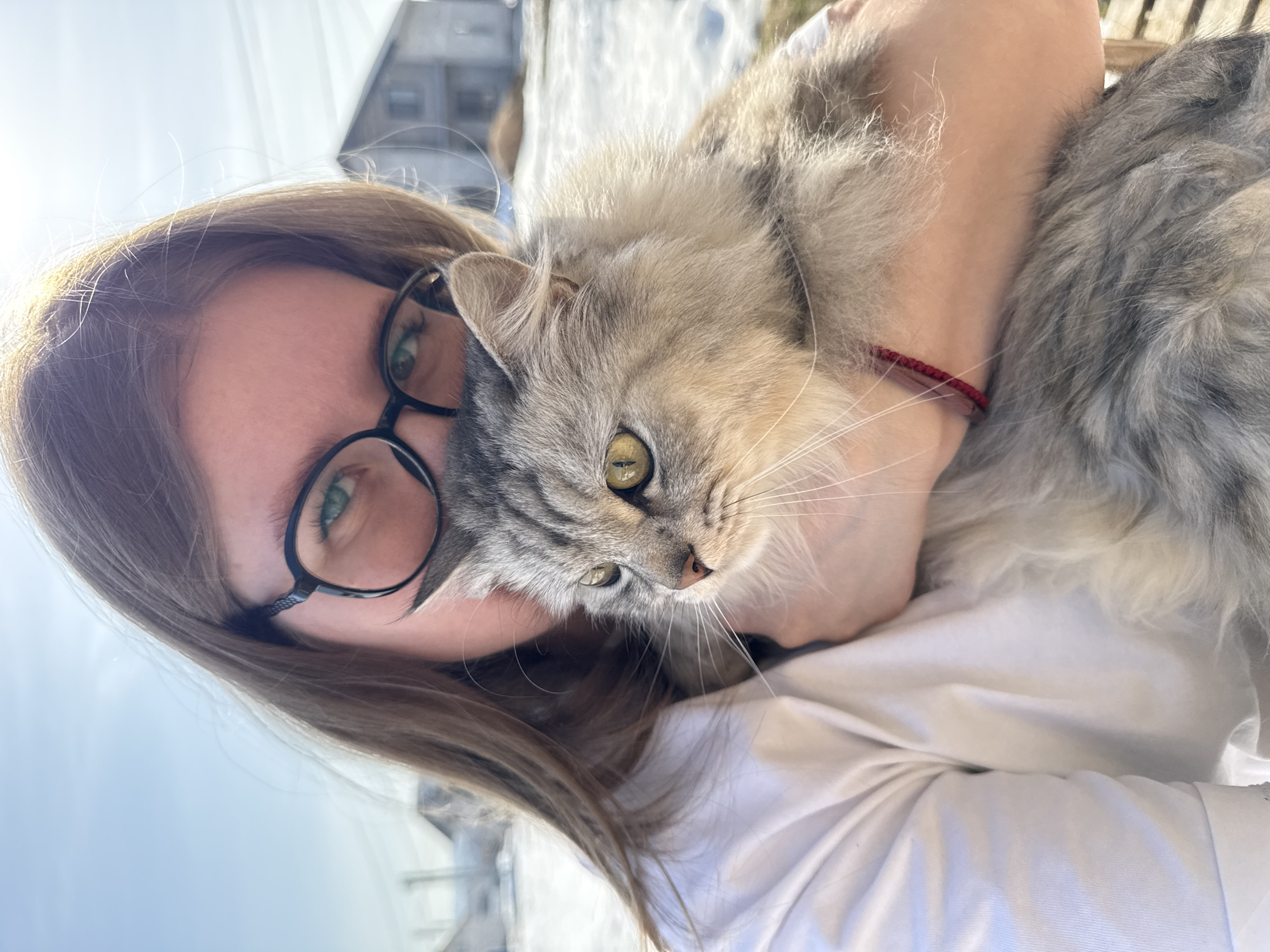 A woman is holding a gray tabby cat in her arms. The woman has light brown hair and is wearing glasses. She is wearing a white shirt. The cat has long fur and yellow-green eyes. The background appears to be outdoors, possibly in a city or town; the image may have been taken in Gornyi (Dvurechenskoe S/P), Russia, although this is not explicitly apparent in the image itself. The woman's arms are wrapped around the cat, suggesting she is holding or hugging it. The focus is on the woman and the cat.