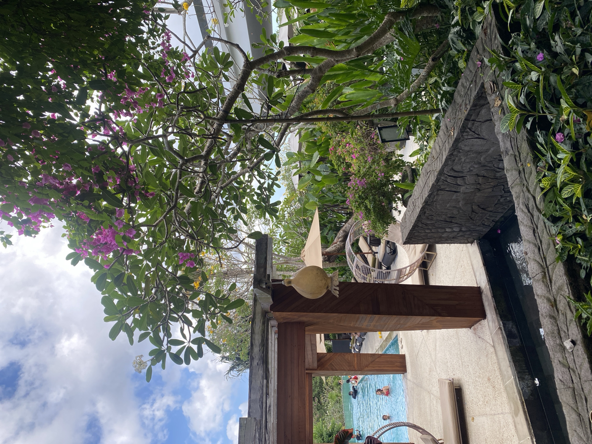 This is an outdoor photograph taken at a resort in Kuta Selatan, Indonesia. The image captures a view of a swimming pool area adjacent to lush tropical vegetation and a partially visible building. In the foreground, there is a stone water feature with a decorative carved edge, and dense green foliage. Beyond this, a paved area surrounds a rectangular swimming pool where several people are visible. Some are swimming, while others are relaxing on loungers. A wooden structure, possibly a pavilion or cabana, with a thatched roof is also present. The background is dominated by the vibrant green of trees and plants, with some purple and yellow flowers adding color. The sky above is blue with scattered white clouds. The overall scene depicts a serene and tropical vacation setting.