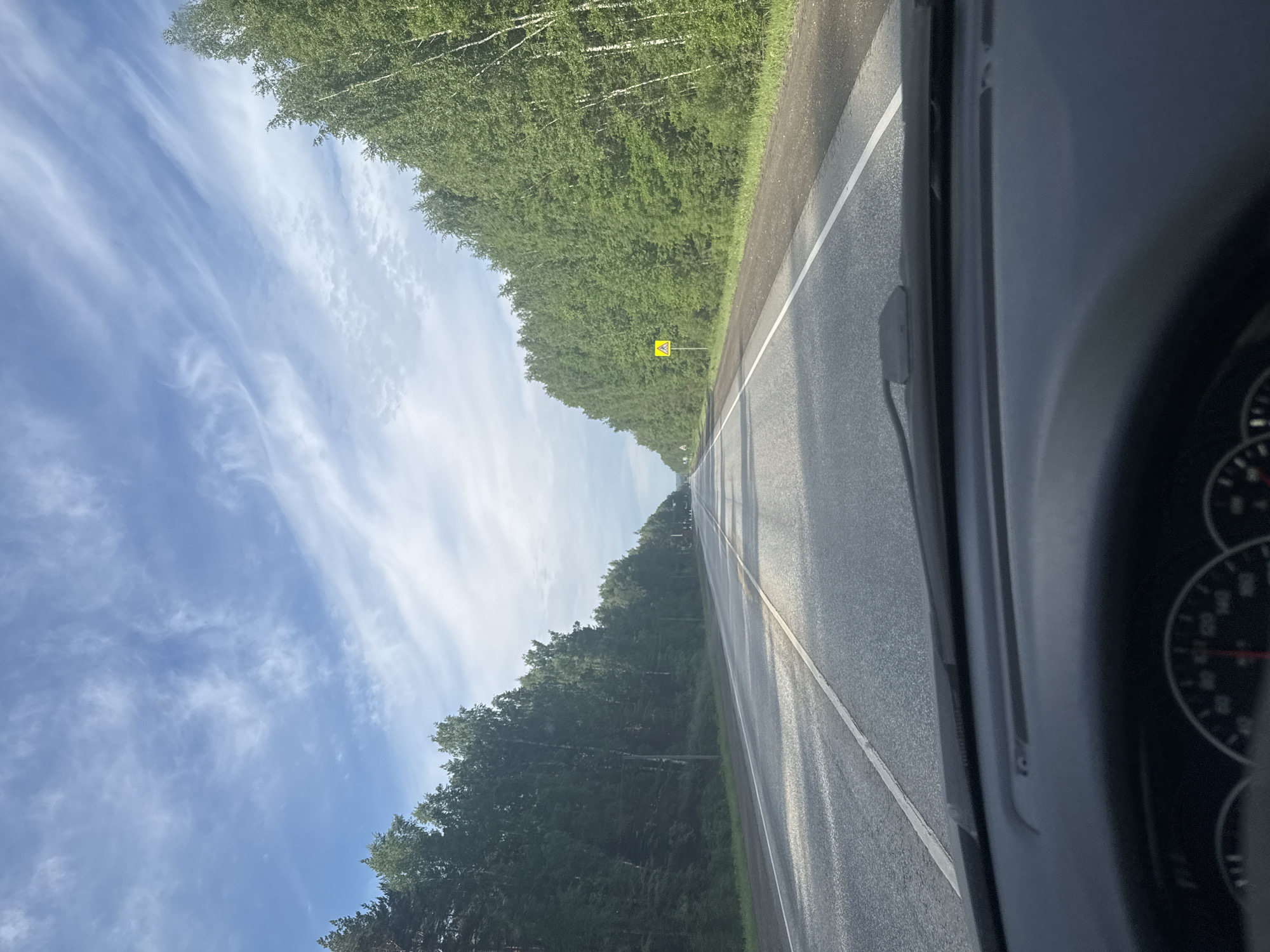 The image captures a first-person perspective from a vehicle traveling on a paved road, likely in the vicinity of Усть-Сыны, Russia. The road, an asphalt surface with a distinct white line marking its edge, extends straight into the horizon. On both sides, dense green forests flank the road, composed primarily of leafy deciduous trees, including some identifiable birches with their characteristic white bark.
Above the tree line, the sky is a vibrant blue, dotted with wispy white clouds that suggest fair weather. Sunlight illuminates the scene, casting subtle shadows across the road surface. The perspective is from inside a car, with the dark dashboard and a portion of the instrument cluster, including a gauge with red markings, visible on the right side of the frame. A part of the windshield wiper mechanism is also discernible.
In the mid-ground, on the left side of the road, a yellow diamond-shaped road sign with black pictograms is visible, serving as a warning or informational sign; based on typical Russian road signs, it likely indicates "Children" or a similar caution.
The scene conveys the experience of driving through a rural, forested landscape on a clear, bright day. There are no people visible, and no readable text other than the indistinguishable symbols on the distant road sign. The overall atmosphere is one of serene travel.