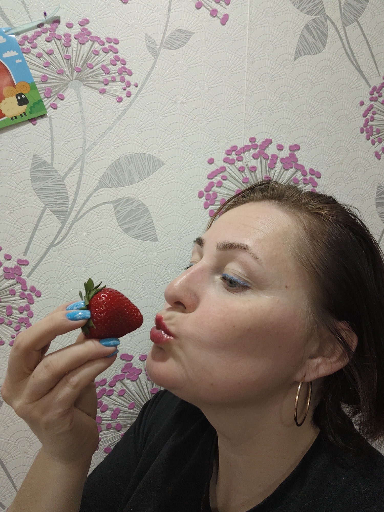 A fair-skinned woman is holding a strawberry in front of her face as if kissing it. Her nails are painted bright blue. She is wearing large, gold hoop earrings. She has auburn hair pulled back from her forehead. She wears bright red lipstick and subtle blue eye makeup. The background is patterned wallpaper with stylized silver leaves and pink flowers. In the upper left corner of the frame, a cartoon sticker of a sheep is partially visible. The location is likely Markovskii, Russia, based on the user's query.