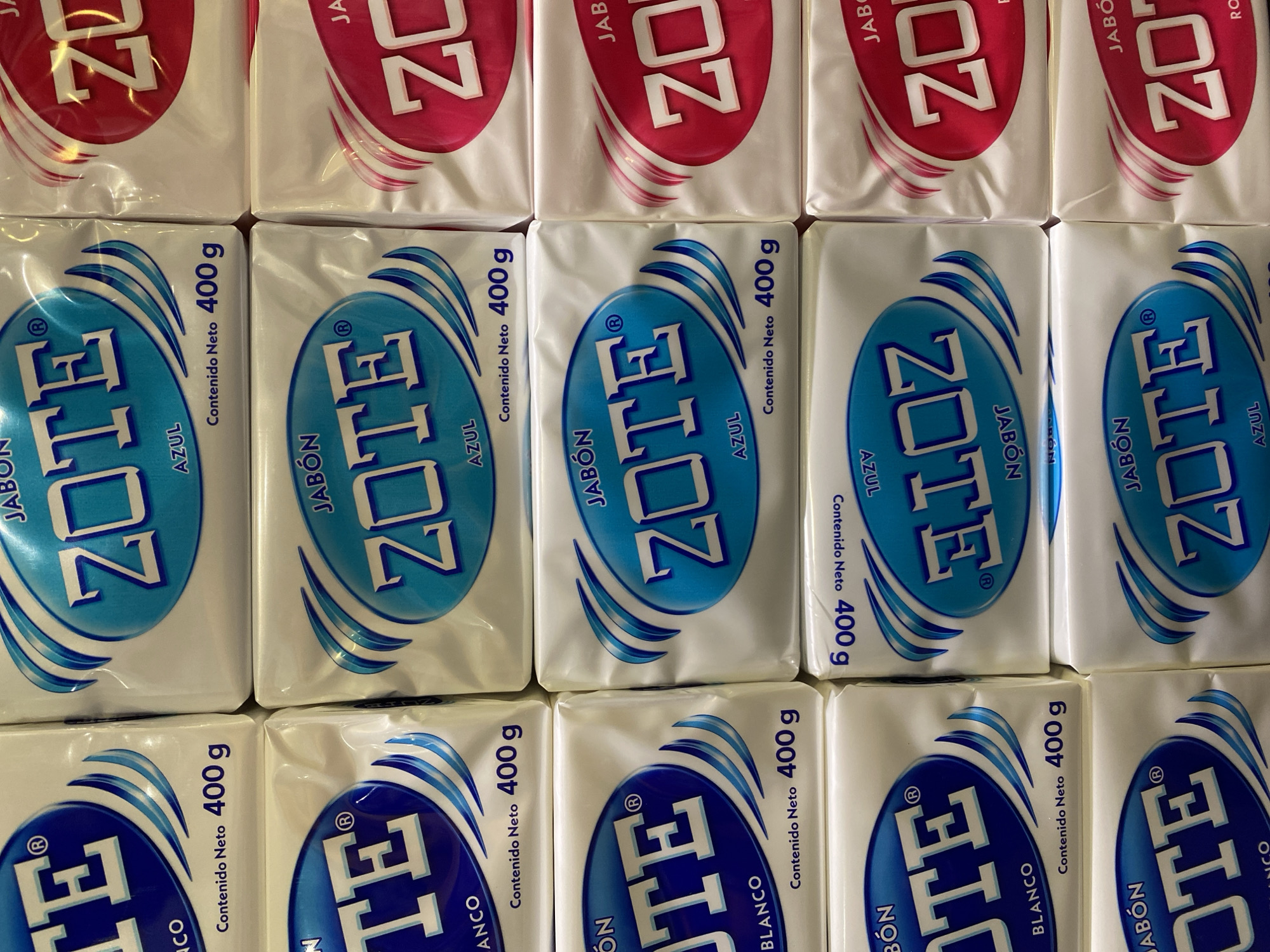 The image shows a close-up view of several bars of ZOTE laundry soap arranged in a grid pattern. The soap bars are predominantly white with different colored oval logos: red, light blue, and dark blue. Each bar's packaging indicates a net weight of 400 g (grams) and the Spanish words "JABÓN" (soap), "AZUL" (blue), and "BLANCO" (white). The brands are ZOTE, AZUL, and BLANCO. There is no information about the location in San Bernardino Tlaxcalancingo, Mexico, or any people in the scene.