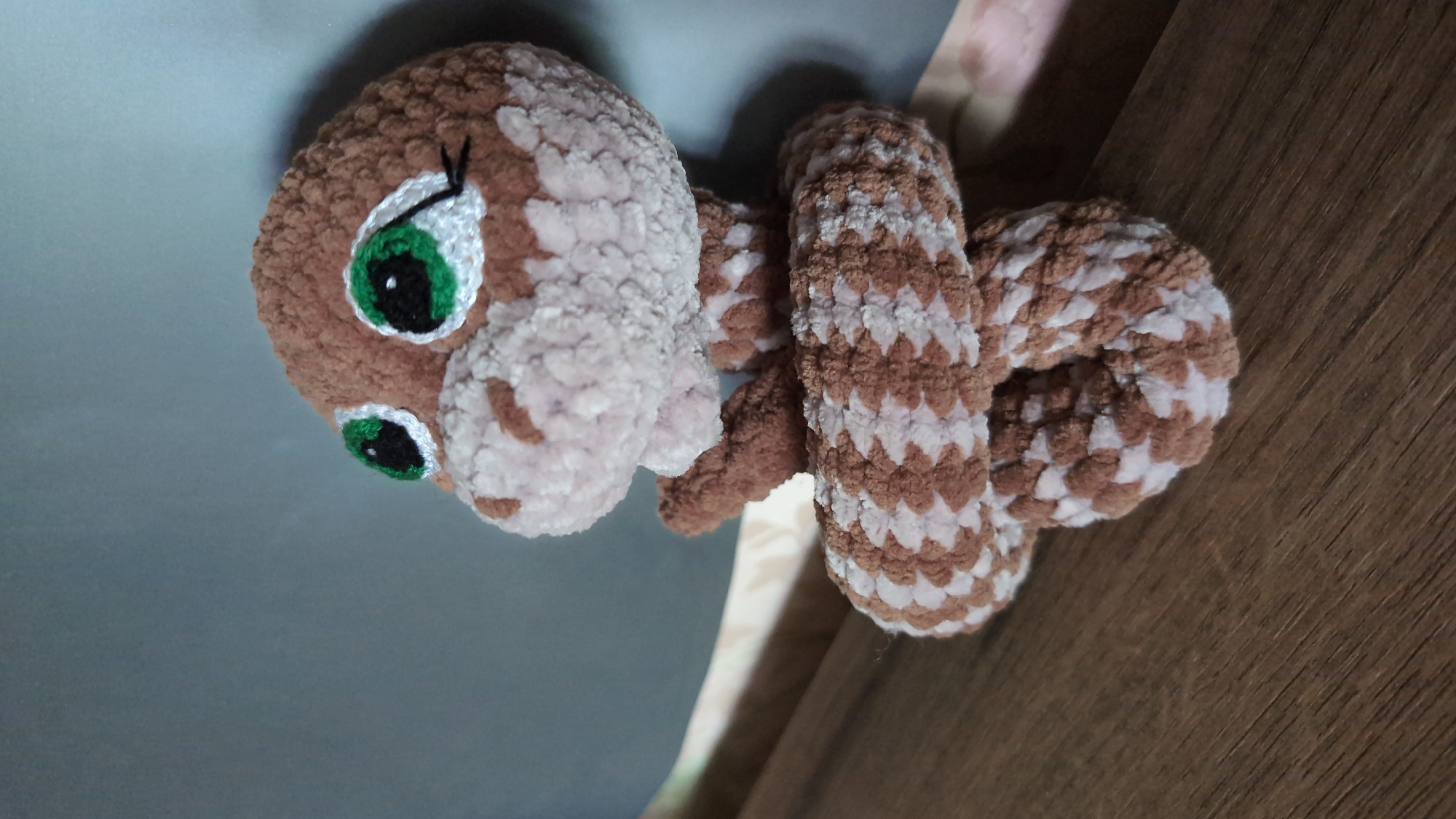 This is a close-up, slightly overhead shot of a crocheted toy snake. The snake has a round head with large, green, expressive eyes outlined in black and white. It has small, dark eyelashes. The head is a light brown color, and the rest of its body is segmented, alternating between brown and white stripes. The snake is coiled, with its body wrapped around a wooden surface, possibly a table or shelf. The background consists of a grey surface on the left and the wooden surface on the right. The lighting suggests an indoor setting, possibly during the day. There is no visible text in the image. The toy appears to be handmade and is positioned as if it is resting or draped over the wooden surface. The overall impression is of a cute and whimsical toy.