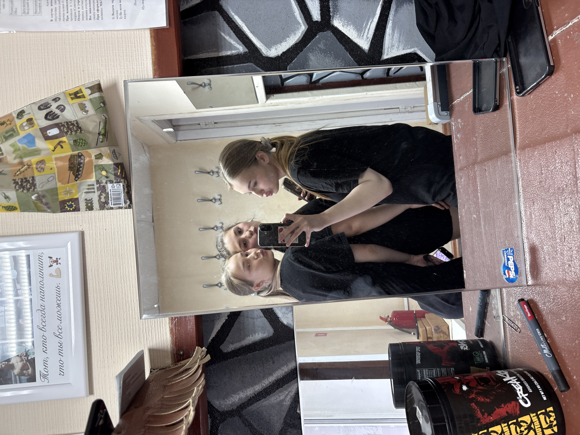 The image shows a rectangular mirror reflecting two young women taking a selfie. The women are dressed in black clothing. One woman is standing slightly behind the other, both holding a smartphone to capture the image. The mirror is positioned in a room with a light beige wall and a decorative, possibly hand-painted, mural with abstract black and grey patterns. Adjacent to the mirror, there is a framed picture with text in Russian, a decorative object resembling a wing, and a colorful item with military-themed designs. On the surface next to the mirror are several objects, including a black phone, two black containers labeled "CREAK," a marker, and a "Pepsi" sticker. The image appears to be taken in a domestic setting, likely in the city of Chaikovsky, Russia, based on the Russian text.