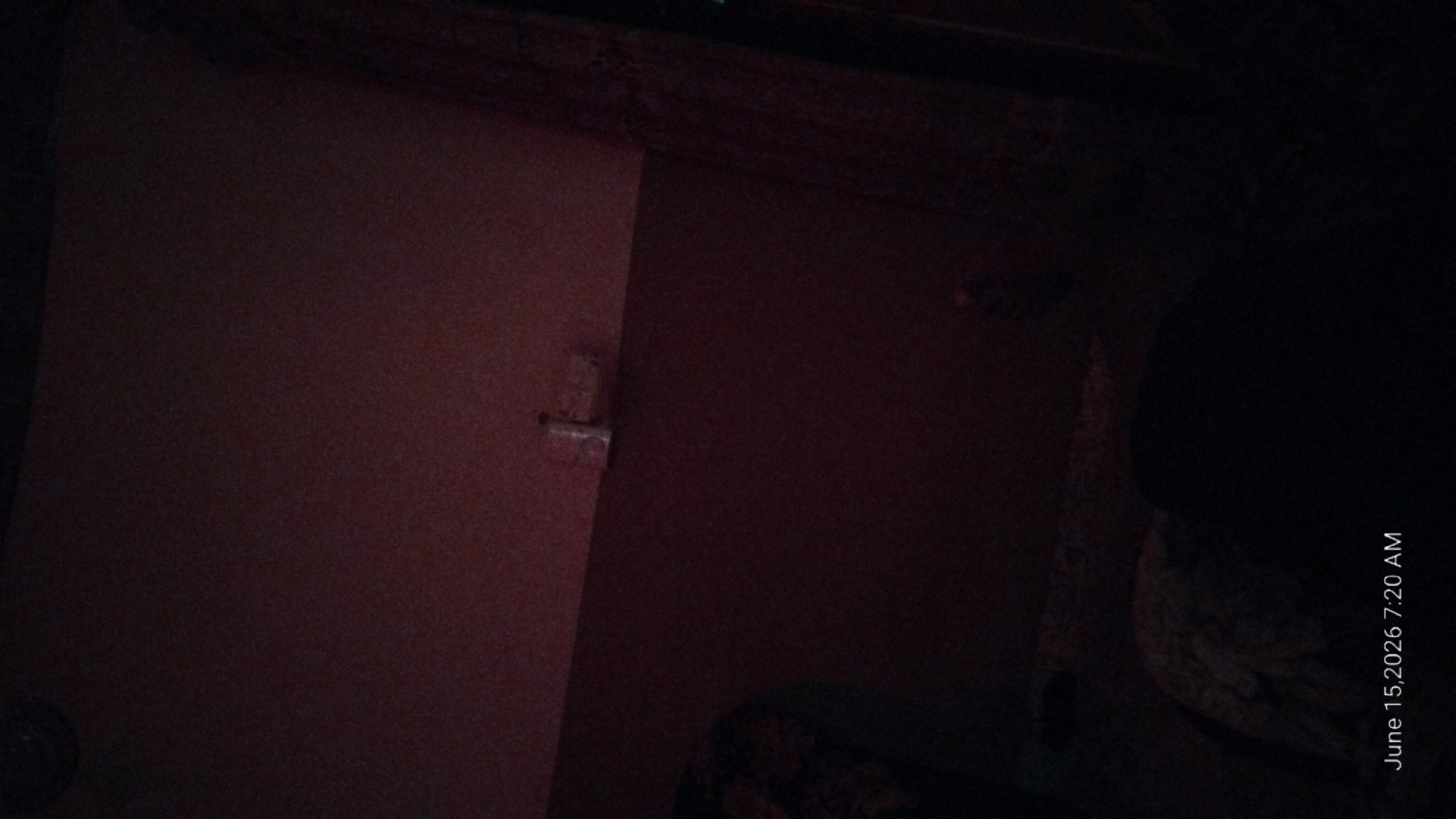 The image is a dimly lit indoor scene, likely a bedroom, with a textured, reddish-brown wall. A white electrical outlet or light switch is visible on the wall towards the upper right. In the foreground, a person's legs and feet are visible, partially covered by a patterned fabric, suggesting they are lying down. The bottom left corner displays a timestamp: "June 15, 2026 7:20 AM." The overall ambiance is dark and subdued, with no clear indication of the city of Howayi, Nigeria.