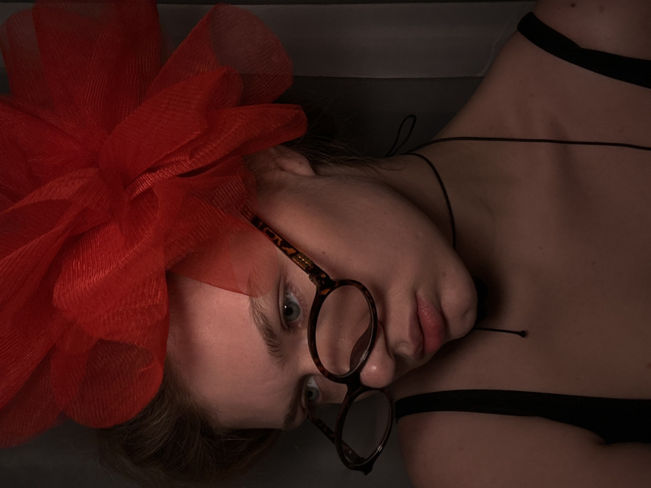 This close-up portrait features a female individual, likely indoors. The specific location within Kazan, Russia, cannot be determined from the image, which displays a plain, dark fabric background, possibly a curtain. The individual possesses light-toned skin, light blue eyes, and light-colored hair pulled back. She wears a large, prominent red headpiece made of sheer, gathered fabric positioned on the right side of her head. Tortoise-shell patterned eyeglasses rest on her nose, tilted downwards, partially obscuring her lower eyelids. A thin black choker with a hanging string is around her neck, and she wears a black strap top. Her gaze is directed downwards and slightly to her left, with a neutral facial expression. Directional lighting illuminates her face and left shoulder, casting shadows on the right side of her face and the background.