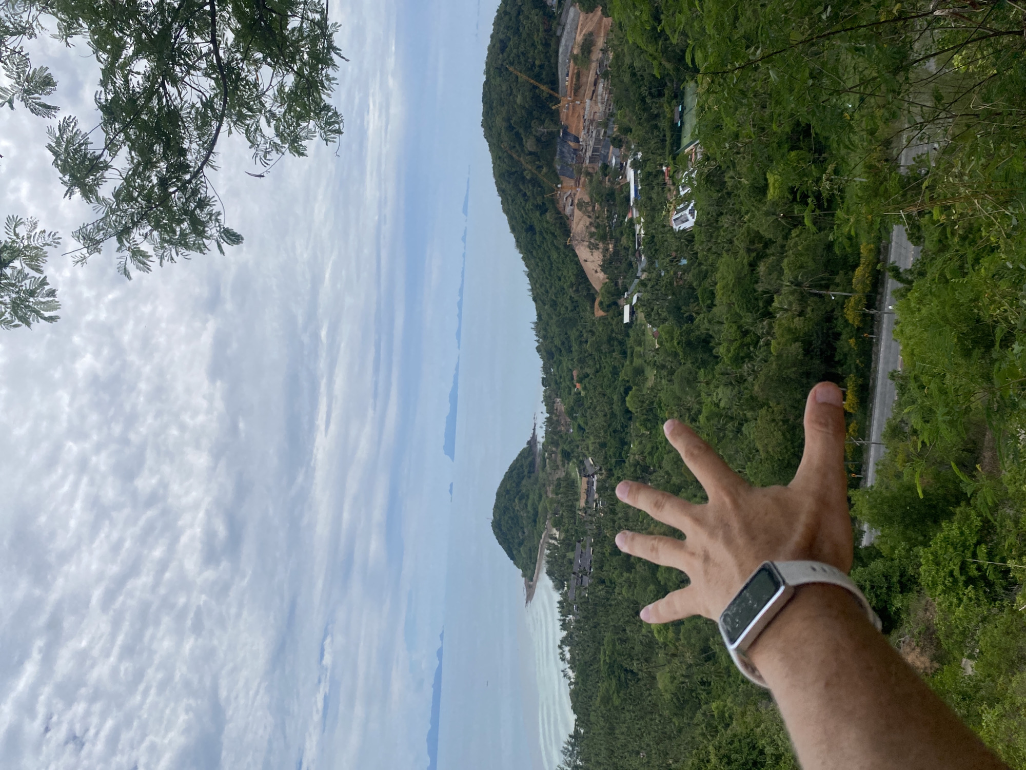 A person's left hand is held out in the foreground, with fingers splayed. The person is wearing a white fitness tracker on their wrist. The background reveals a lush, green landscape of tropical trees and vegetation. In the middle ground, there is a body of water, with a small, tree-covered island situated in the bay. Further inland, a hillside is undergoing significant construction, with cranes and unfinished buildings visible. In the distance, more islands can be seen on the horizon under a cloudy sky. The scene appears to be a scenic overlook in or near Baan Thong Po, Thailand, likely offering a view of the coast and surrounding natural beauty.
