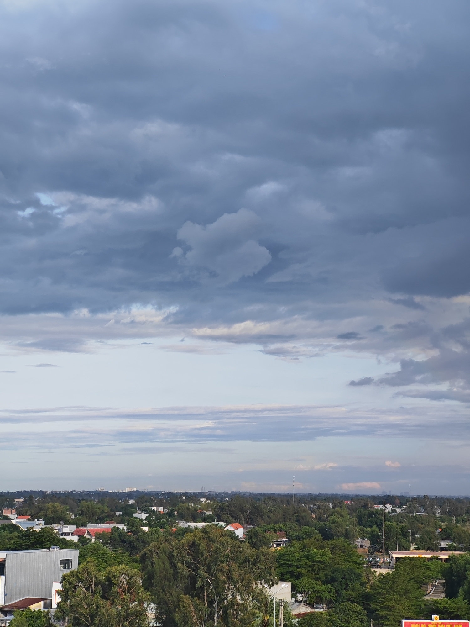 The image captures a panoramic view of a city, likely Đà Nẵng, Vietnam, under a dramatic sky filled with dark, heavy clouds. In the foreground, a building with a corrugated metal facade is visible, partially obscured by lush green trees. The middle ground reveals a dense canopy of trees interspersed with residential rooftops and a few larger buildings. A road with streetlights runs through the area, and a few vehicles and a small group of people are discernible near a building with a yellow facade. The background shows a flat landscape extending towards a hazy horizon, with distant buildings and what appear to be communication towers. The overall atmosphere is overcast and potentially indicative of approaching rain.