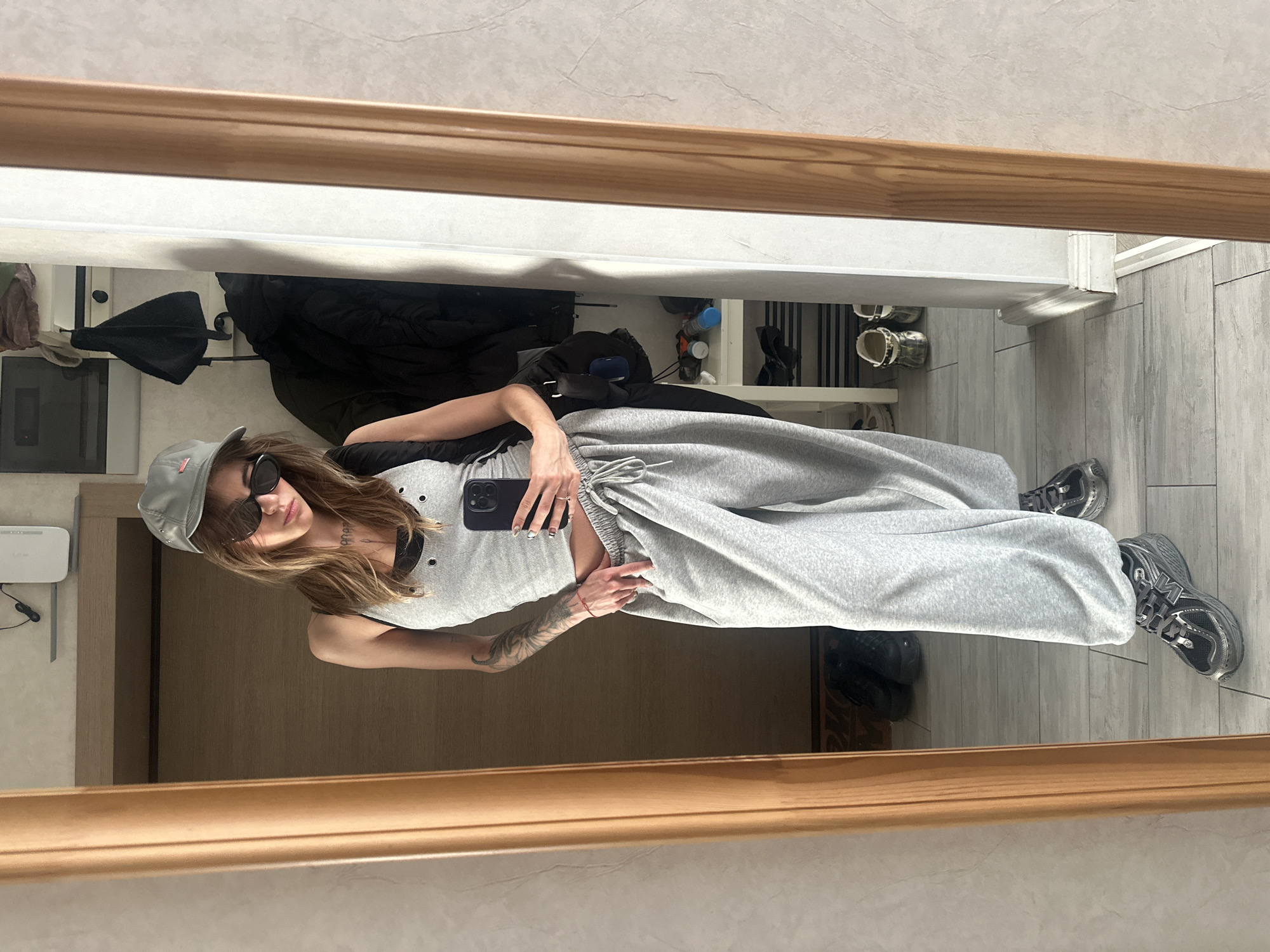 The image shows a young woman posing for a selfie in a mirror. She is wearing a grey tank top, grey sweatpants, a grey baseball cap, and sunglasses. She is also carrying a black backpack. The background appears to be an indoor space, possibly a home or a hotel room, with a wooden door and a white wall. A shelf with various items is visible in the reflection of the mirror.