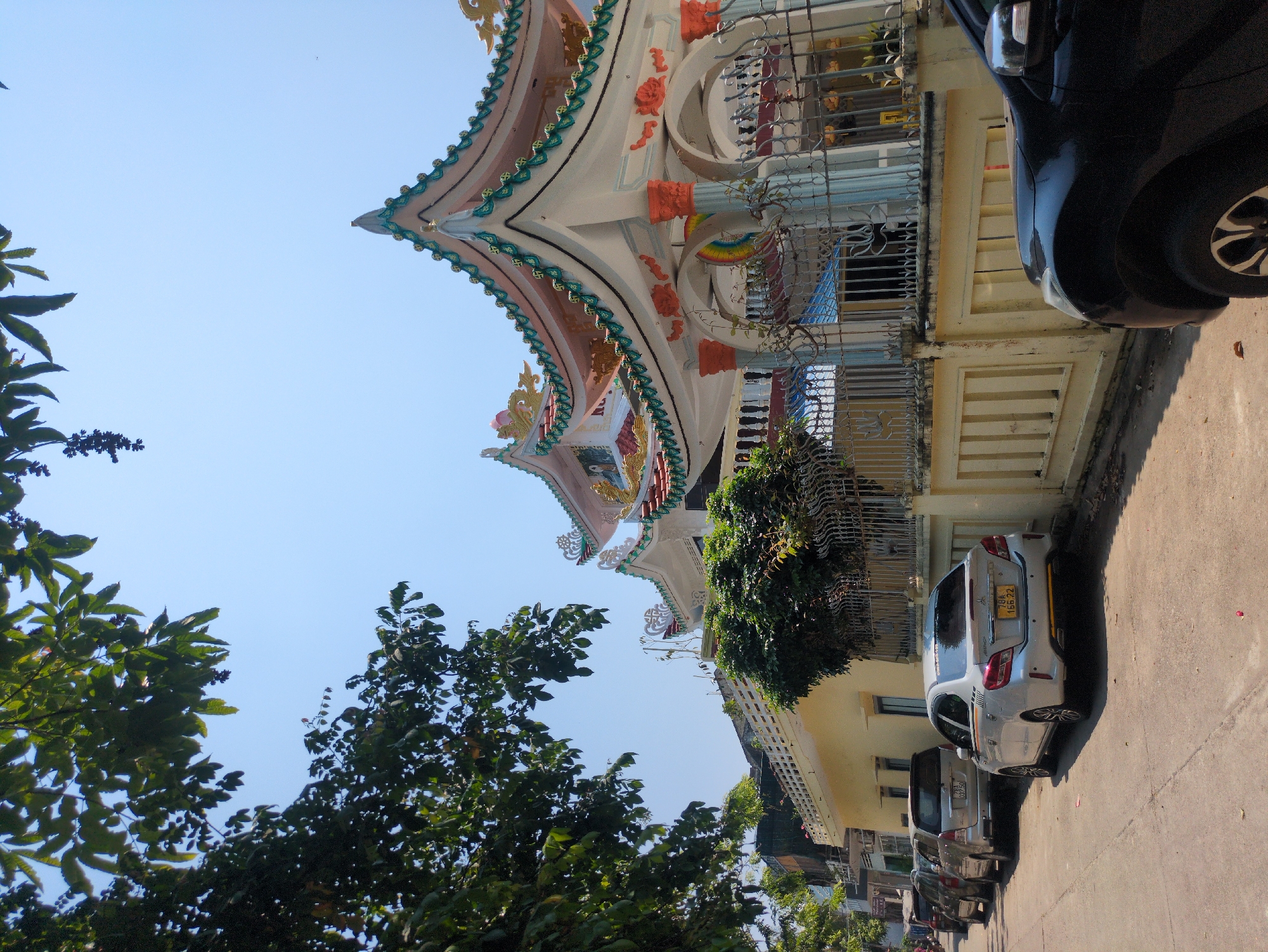 A street scene in Tuy Hòa, Vietnam, captures a building with elaborate, tiered eaves adorned with decorative carvings. The building's facade, painted in light yellow, features grilled balconies and is adjacent to a narrow street lined with parked cars, including a silver sedan and a black SUV. Green trees frame the view, with a clear blue sky overhead.