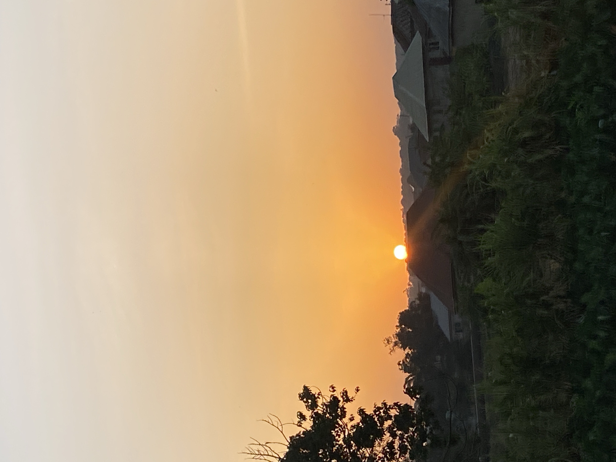 An outdoor, angled eye-level shot shows a sunset over Sabon Tasha, Nigeria. The orange sun is positioned in the middle, and to the right are trees and the roofs of houses. A clear sky fills the left side of the photo.
